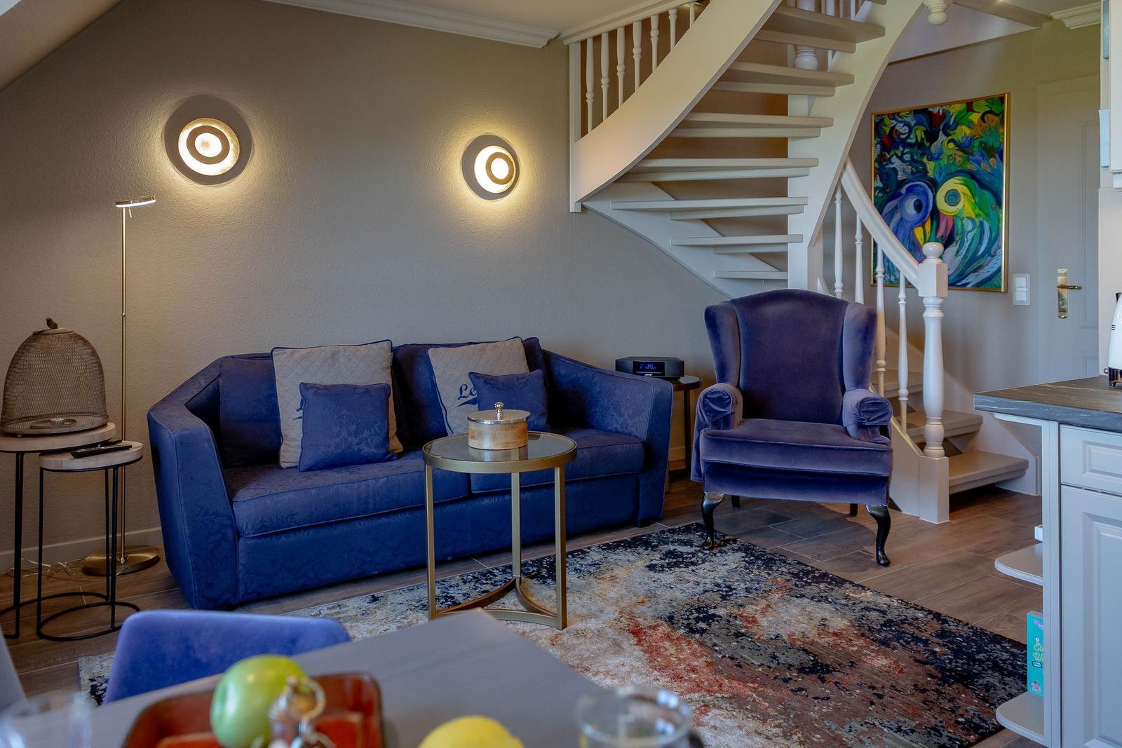 Wohnbereich mit blauem Sofa, Sessel und Treppe. Wandlampen und Teppich sind sichtbar.