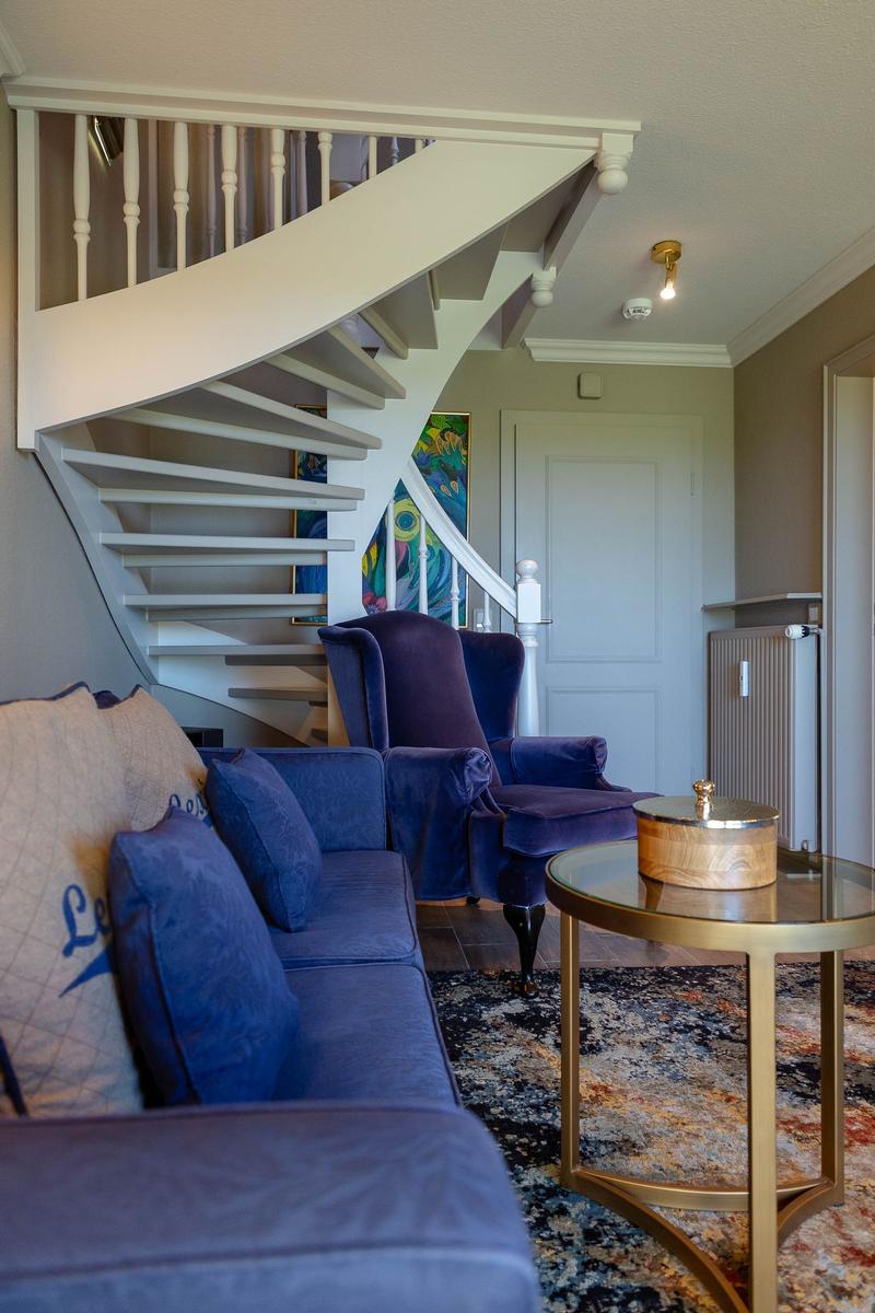 Ein Wohnzimmer mit blauem Sofa, Sessel und Spiraltreppe. Tisch und Teppich sind sichtbar.
