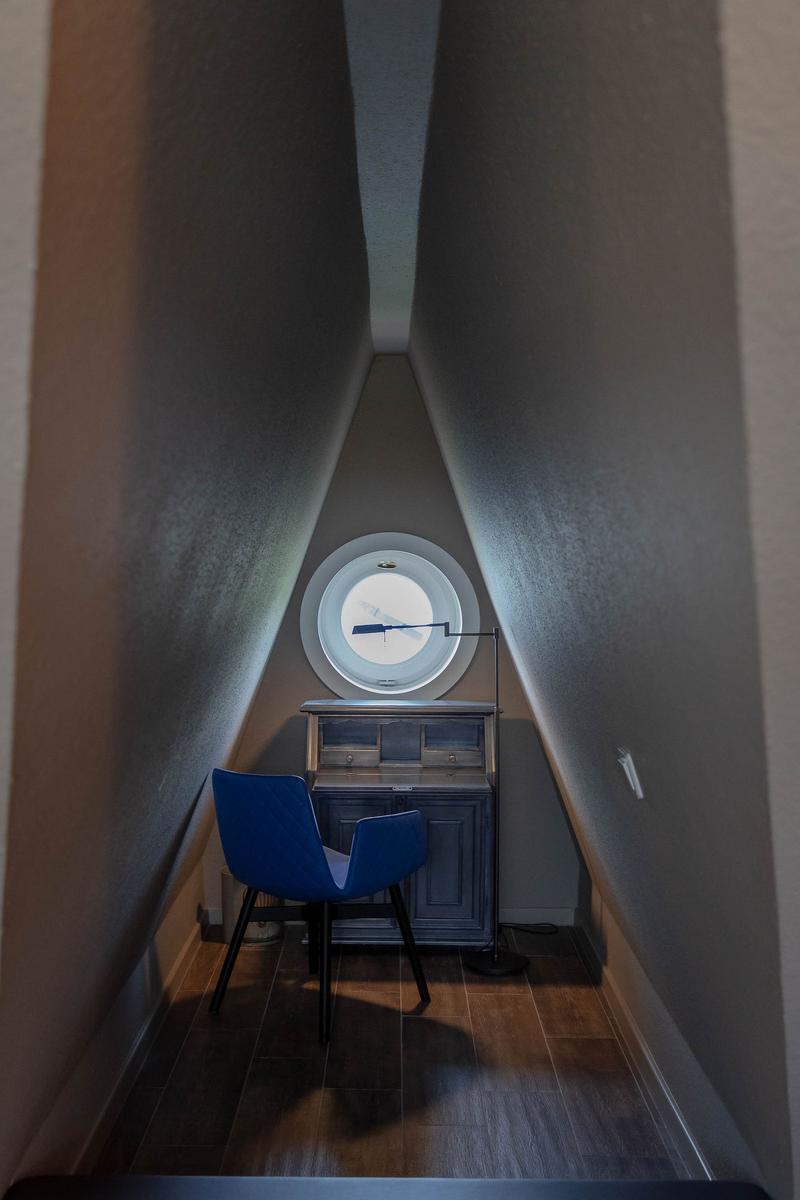 Ein kleiner Raum mit schrägen Wänden, einem runden Fenster und einem blauen Stuhl vor einem Schrank.