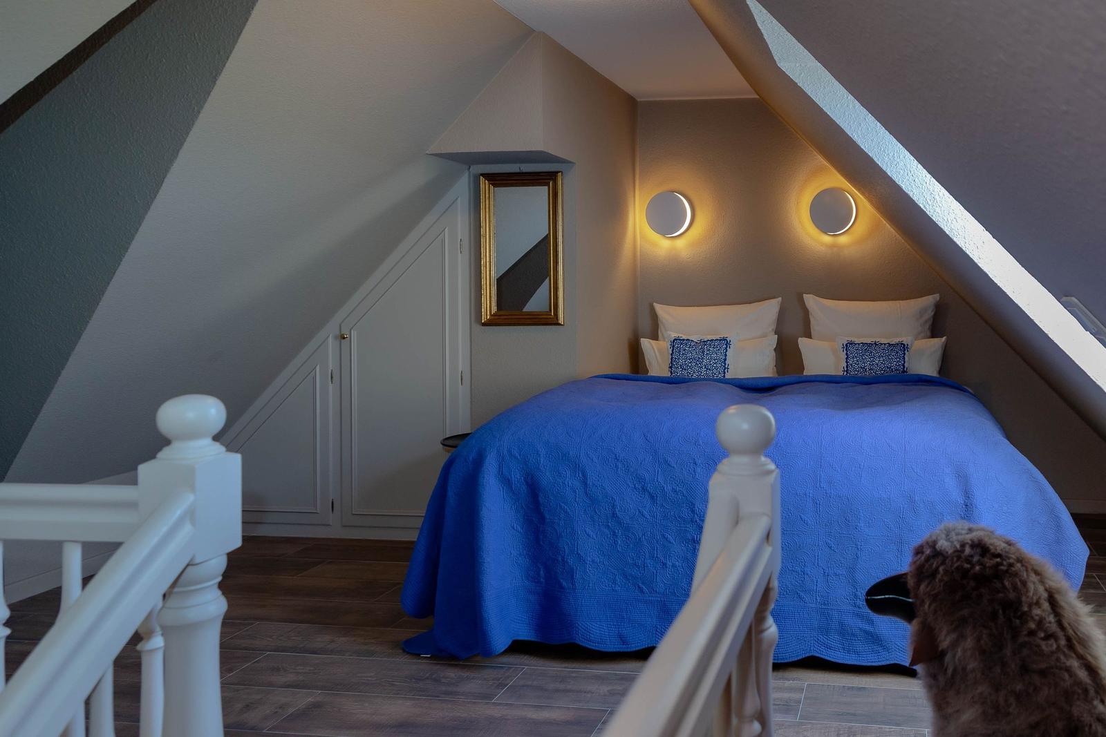 Schlafzimmer mit blauer Bettdecke, zwei runden Wandlampen und einem Spiegel an der Wand.