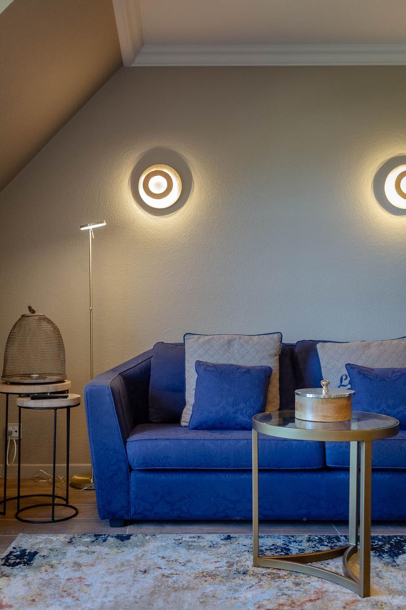 Blauer Sofa mit Kissen, runde Tische und Wandlampen in einem Raum mit schräger Decke.