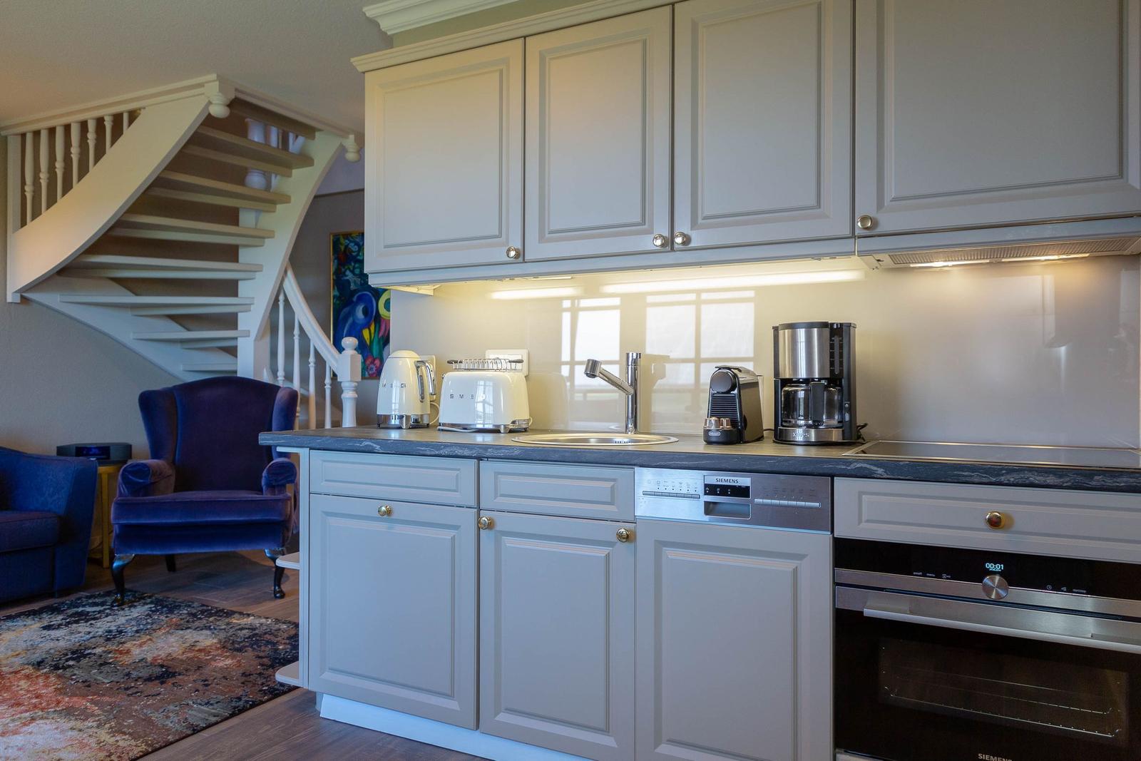 Küche mit hellen Schränken, Edelstahlgeräten und Blick auf Treppenhaus.