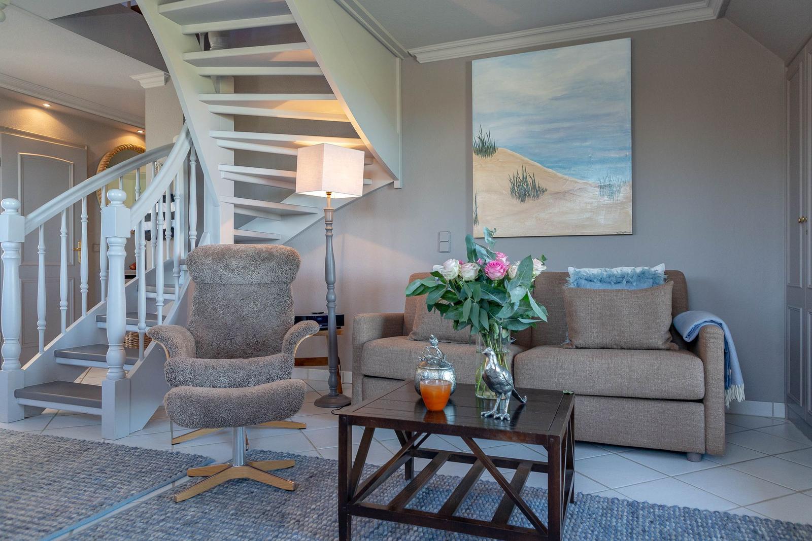 Wohnzimmer mit Sofa, Sessel, Tisch und Treppe. Wandbild zeigt Strand.