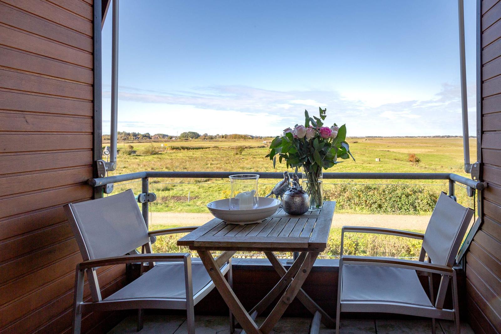 Terrasse mit Tisch, zwei Stühlen und Blumen. Blick auf grünes Feld unter blauem Himmel.