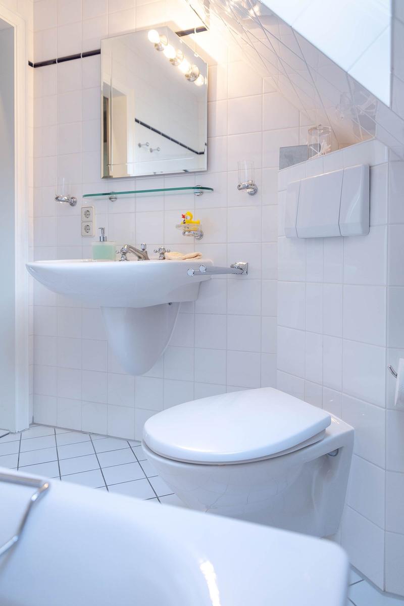 Badezimmer mit Waschbecken, Spiegel und Toilette. Weiße Fliesen und moderne Ausstattung.