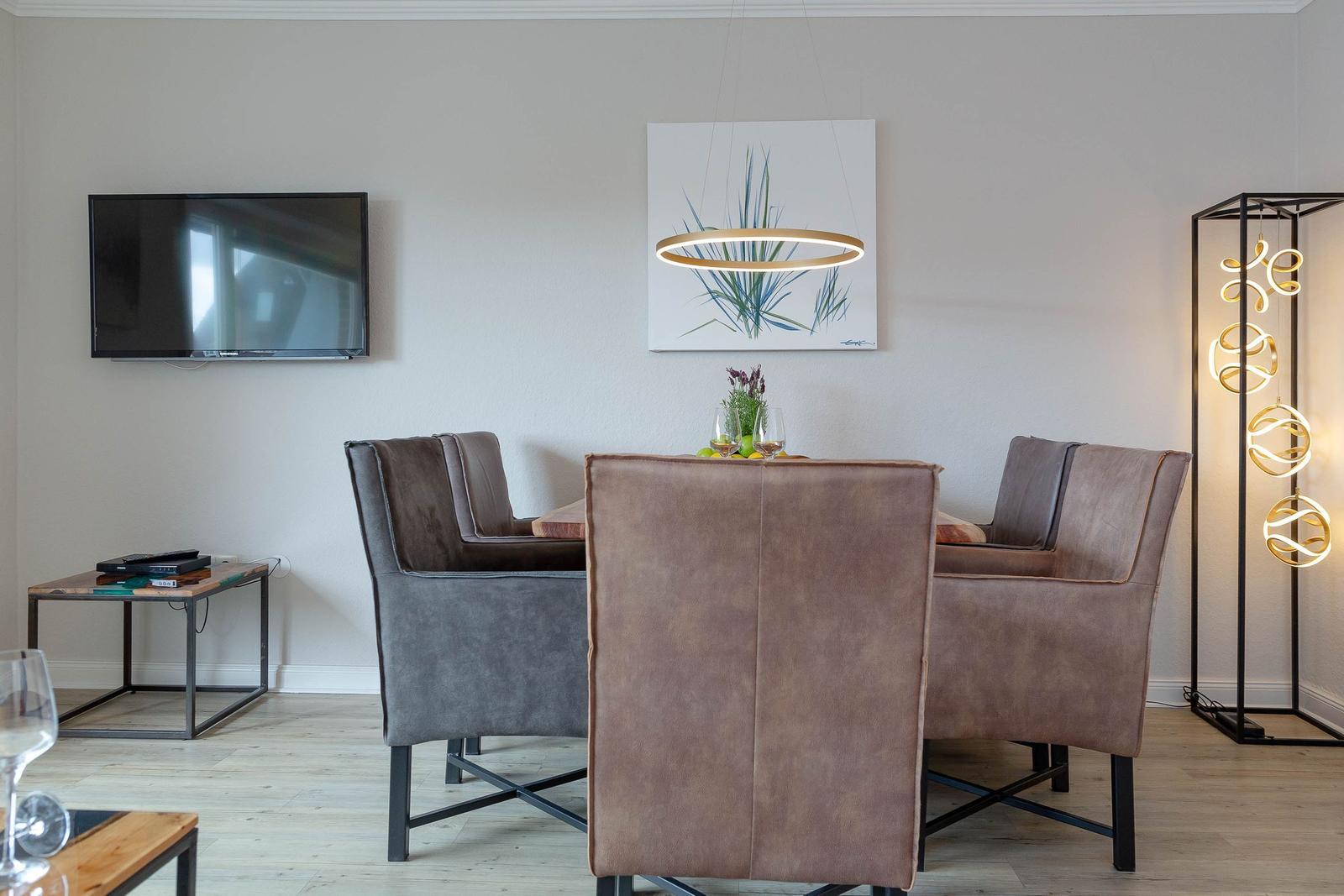 Esszimmer mit Holztisch, vier Stühlen und Wand-TV. Deckenleuchte und Stehlampe sorgen für Beleuchtung.