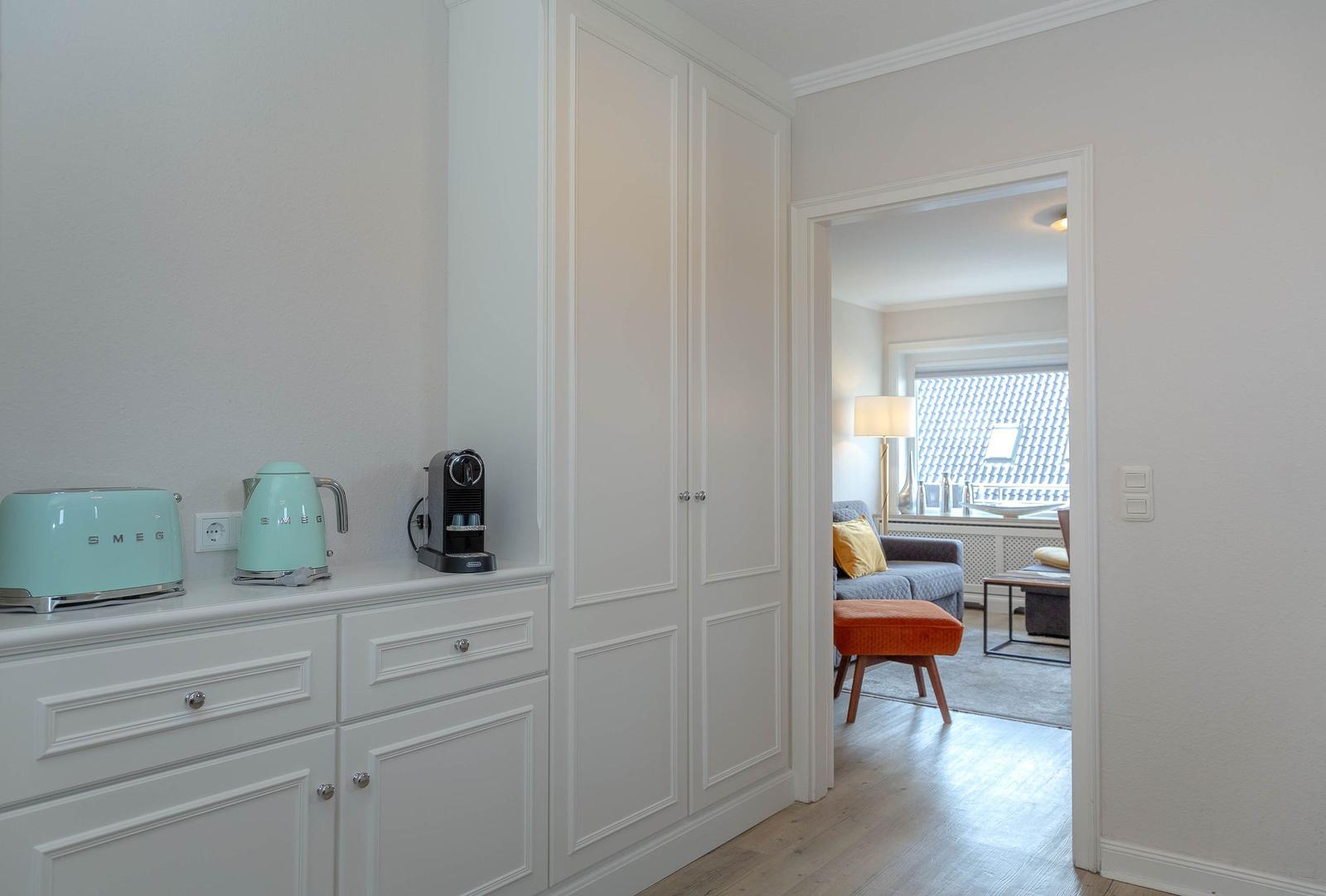 Küchenzeile mit Kaffeemaschine und Kessel. Durchgang in Wohnzimmer mit Sofa und Hocker.