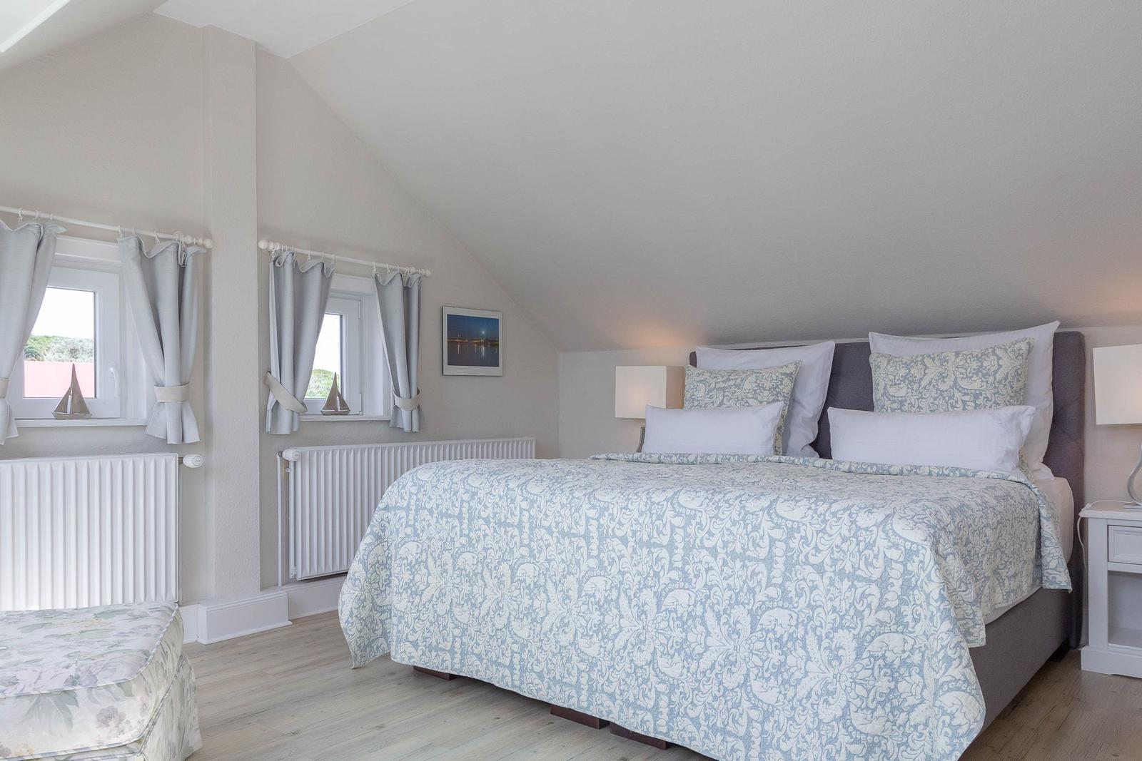 Schlafzimmer mit Doppelbett, zwei Fenstern und Holzboden.