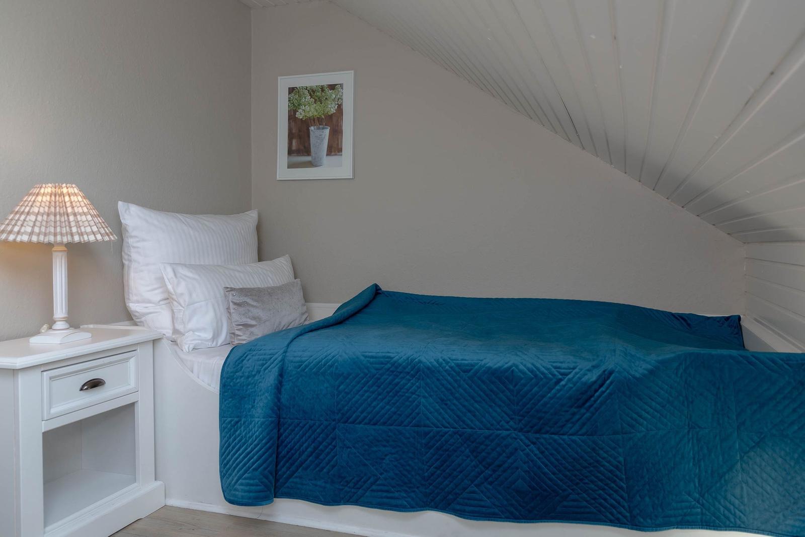 Ein Bett mit blauer Decke, Nachttisch und Wandbild unter schrägem Dach