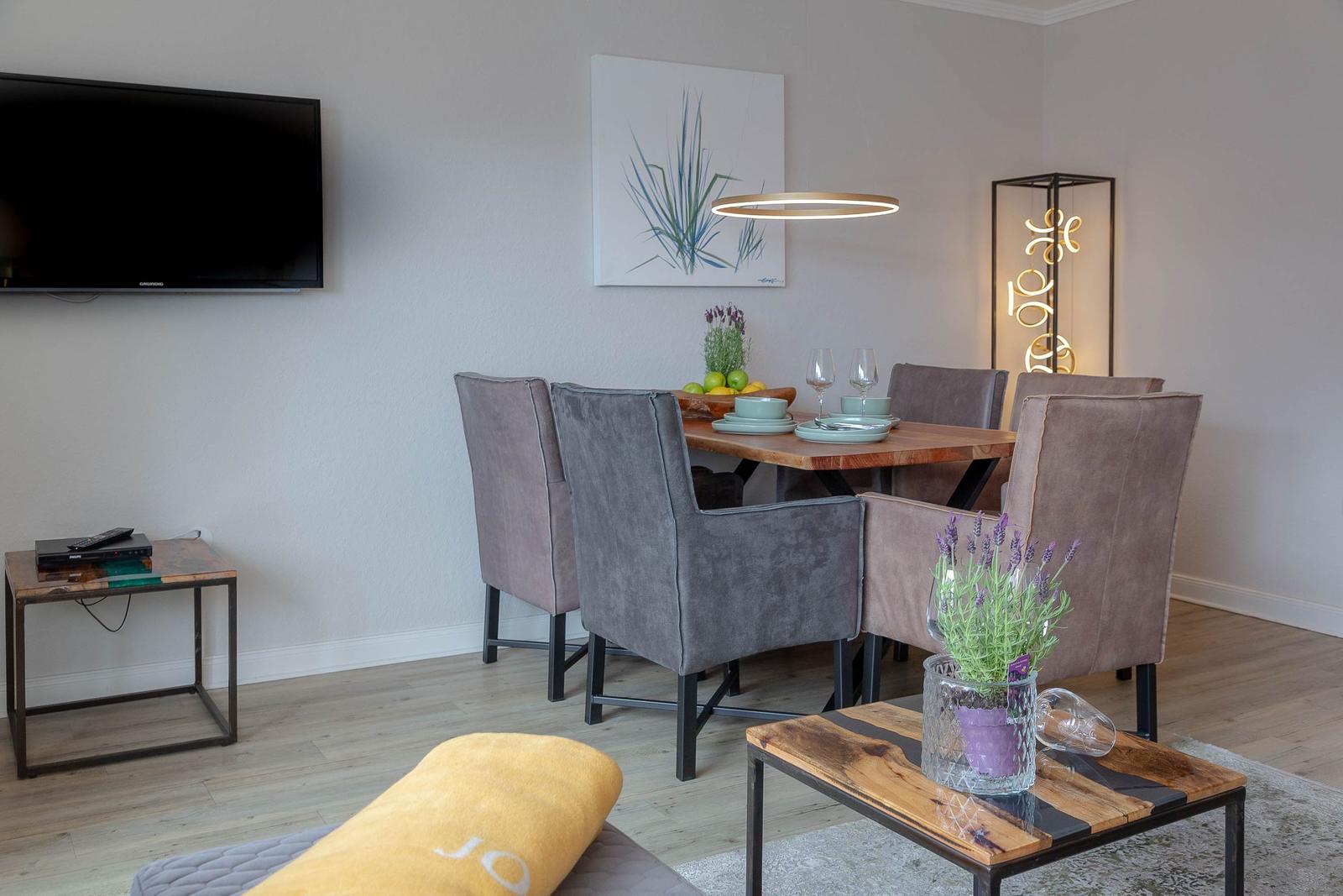 Esszimmer mit Holztisch, grauen Stühlen und Wandbild. Fernseher und Beistelltisch im Vordergrund.