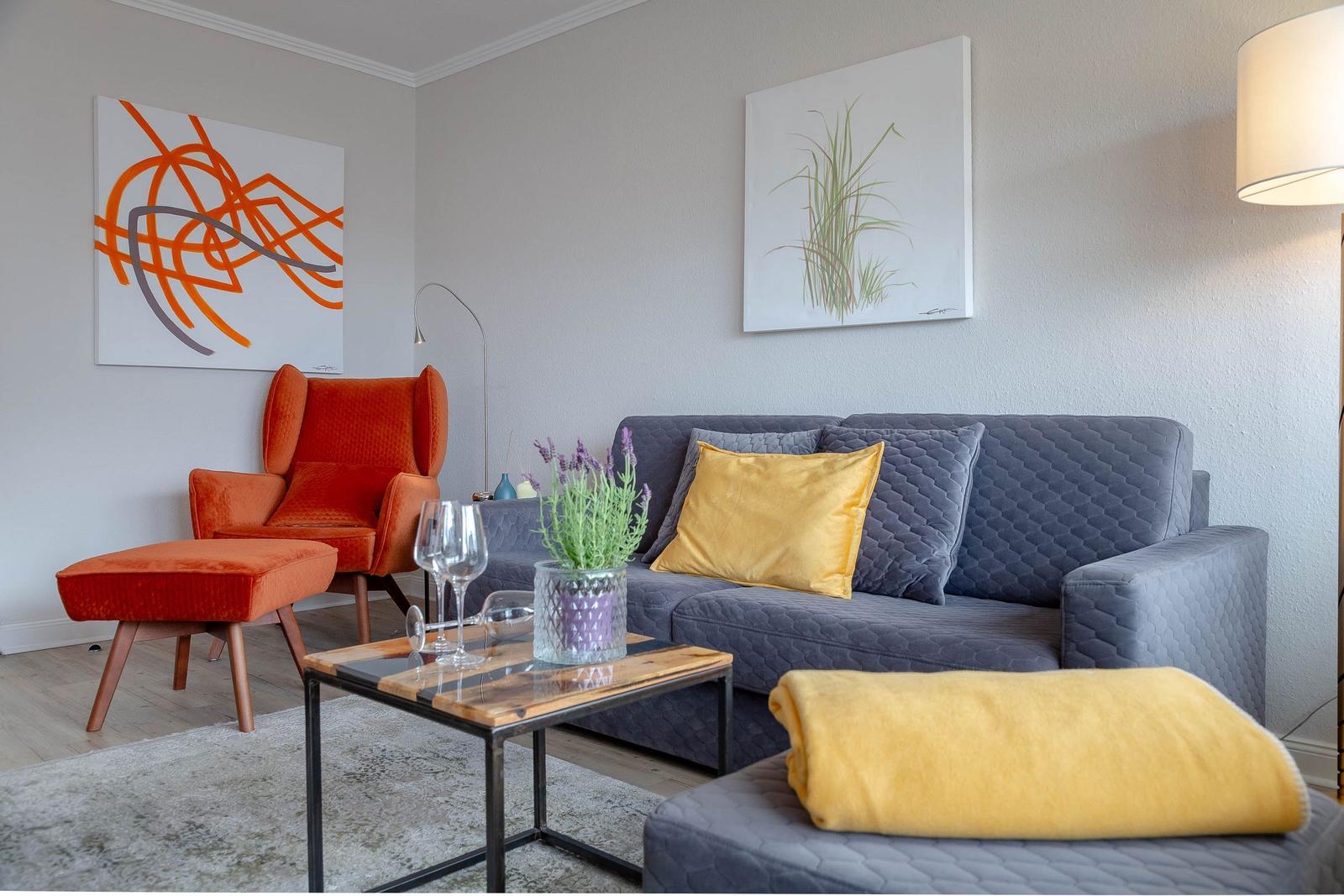 Ein Wohnzimmer mit grauem Sofa, orangefarbenem Sessel und modernen Kunstwerken an den Wänden.