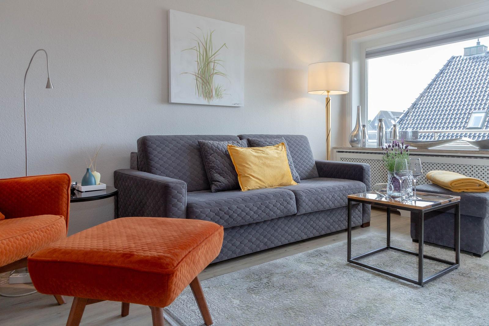 Ein Wohnzimmer mit grauem Sofa, orangefarbenem Sessel und Fensterblick auf Dächer.