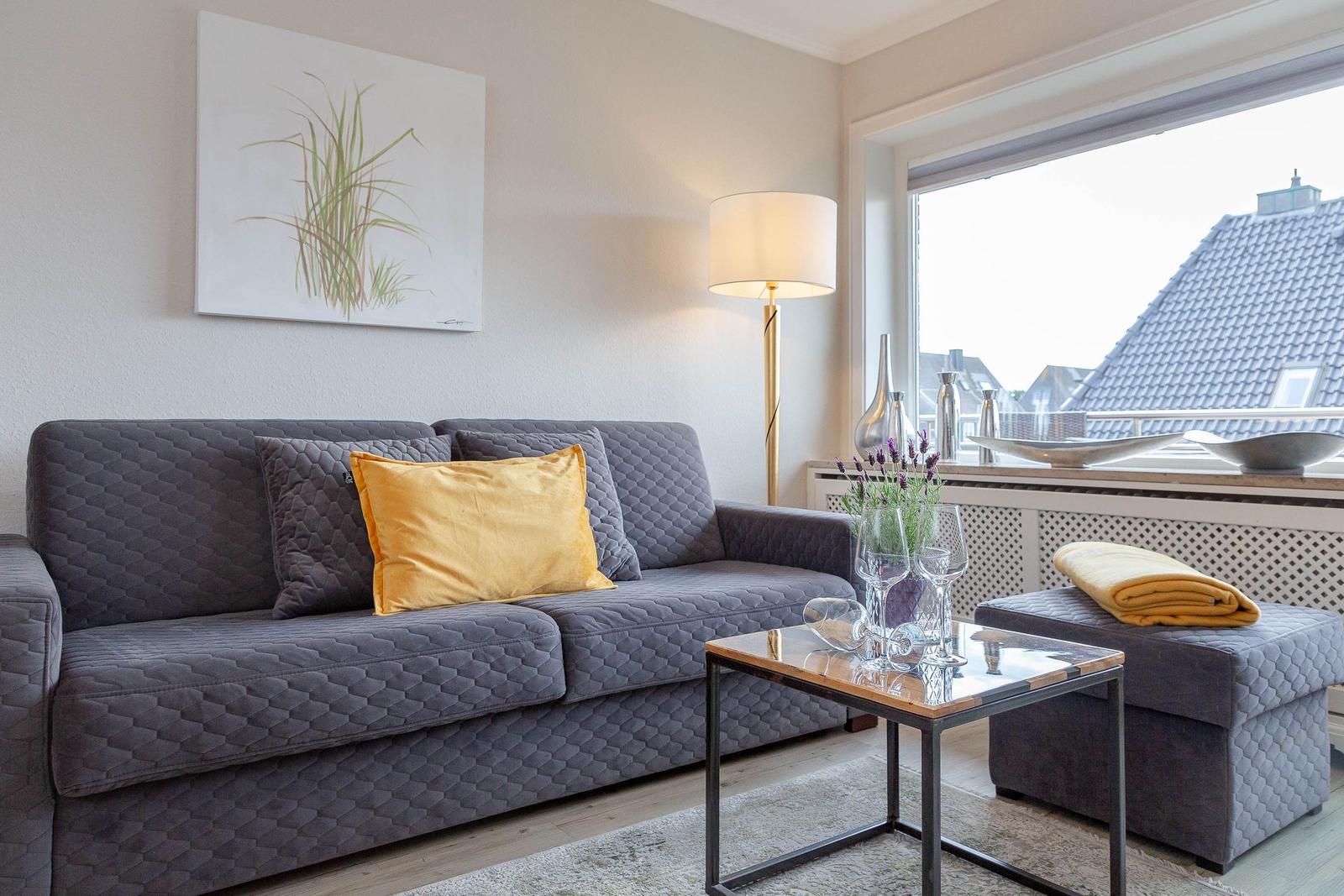 Ein Wohnzimmer mit grauem Sofa, gelben Kissen und einem Fenster mit Blick auf Dächer.