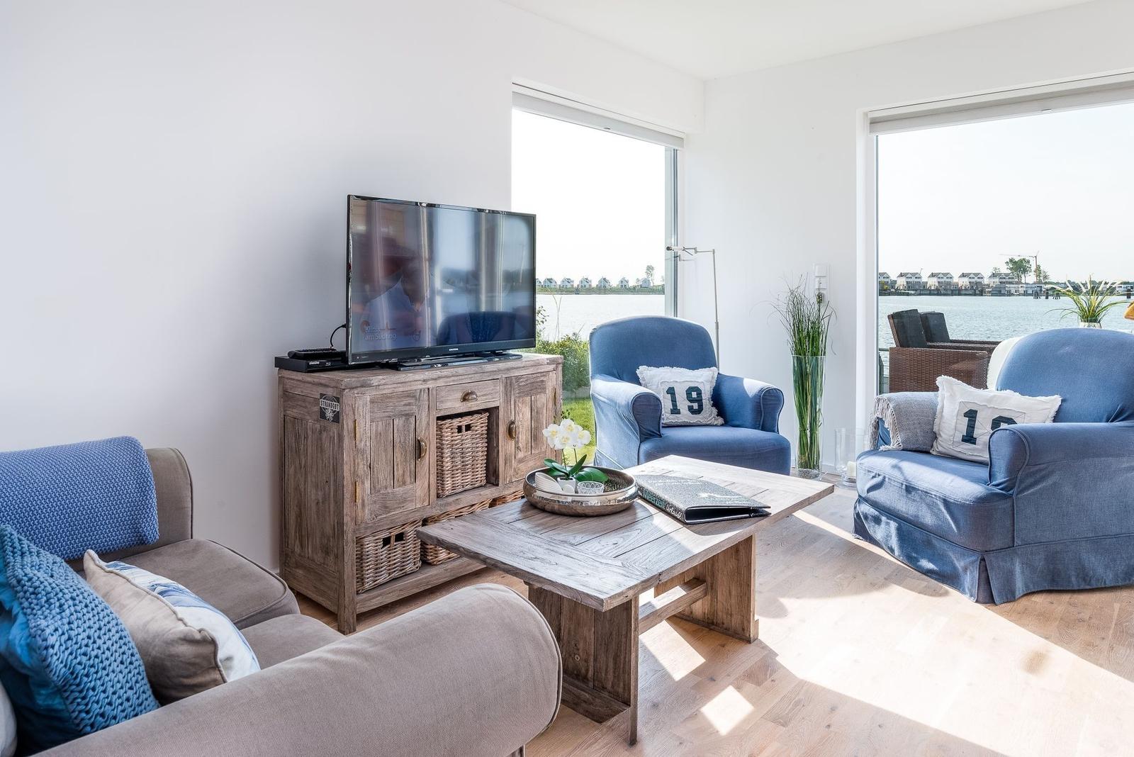 Wohnzimmer mit TV, Holztisch und blauen Sesseln. Blick auf Wasser durch Fenster.