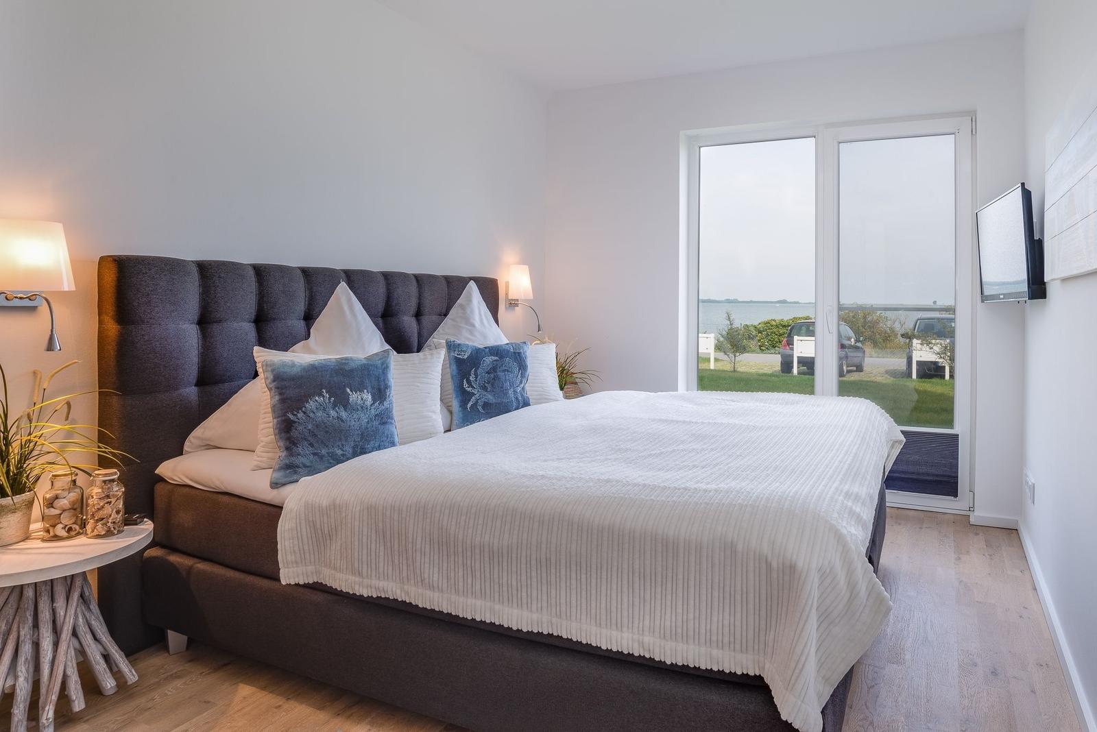 Schlafzimmer mit großem Bett, Fensterblick auf Wasser und Fernseher an der Wand.