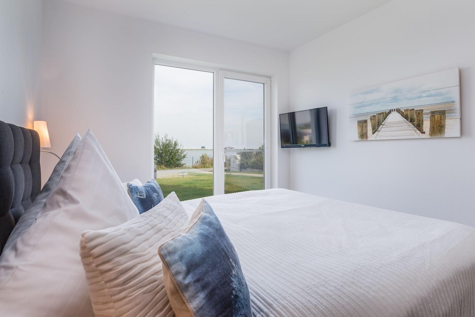 Schlafzimmer mit Bett, TV und Blick auf Grünfläche und Wasser.
