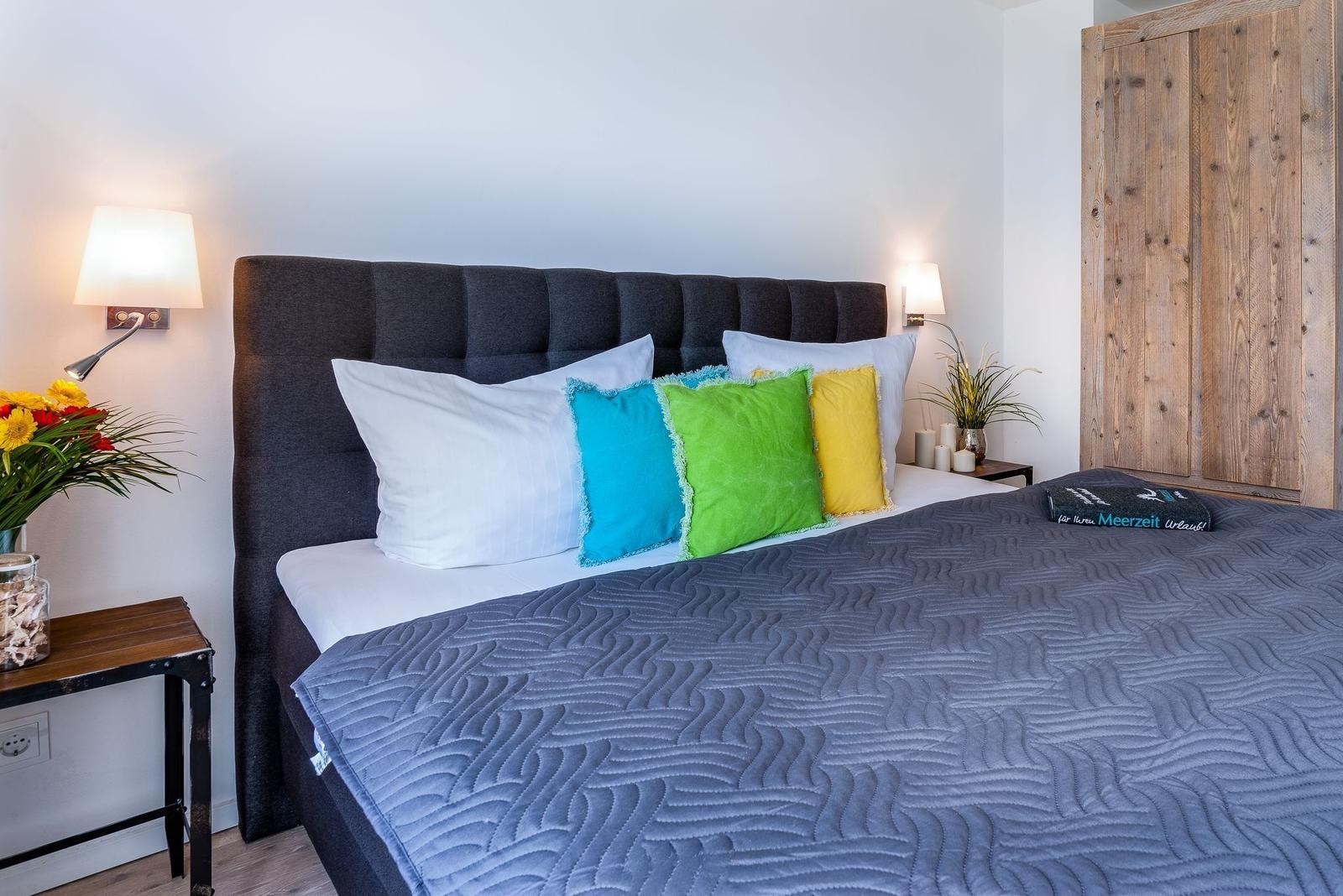Schlafzimmer mit Bett, Kissen und Holzschrank. Beleuchtung und Blumen sind sichtbar.