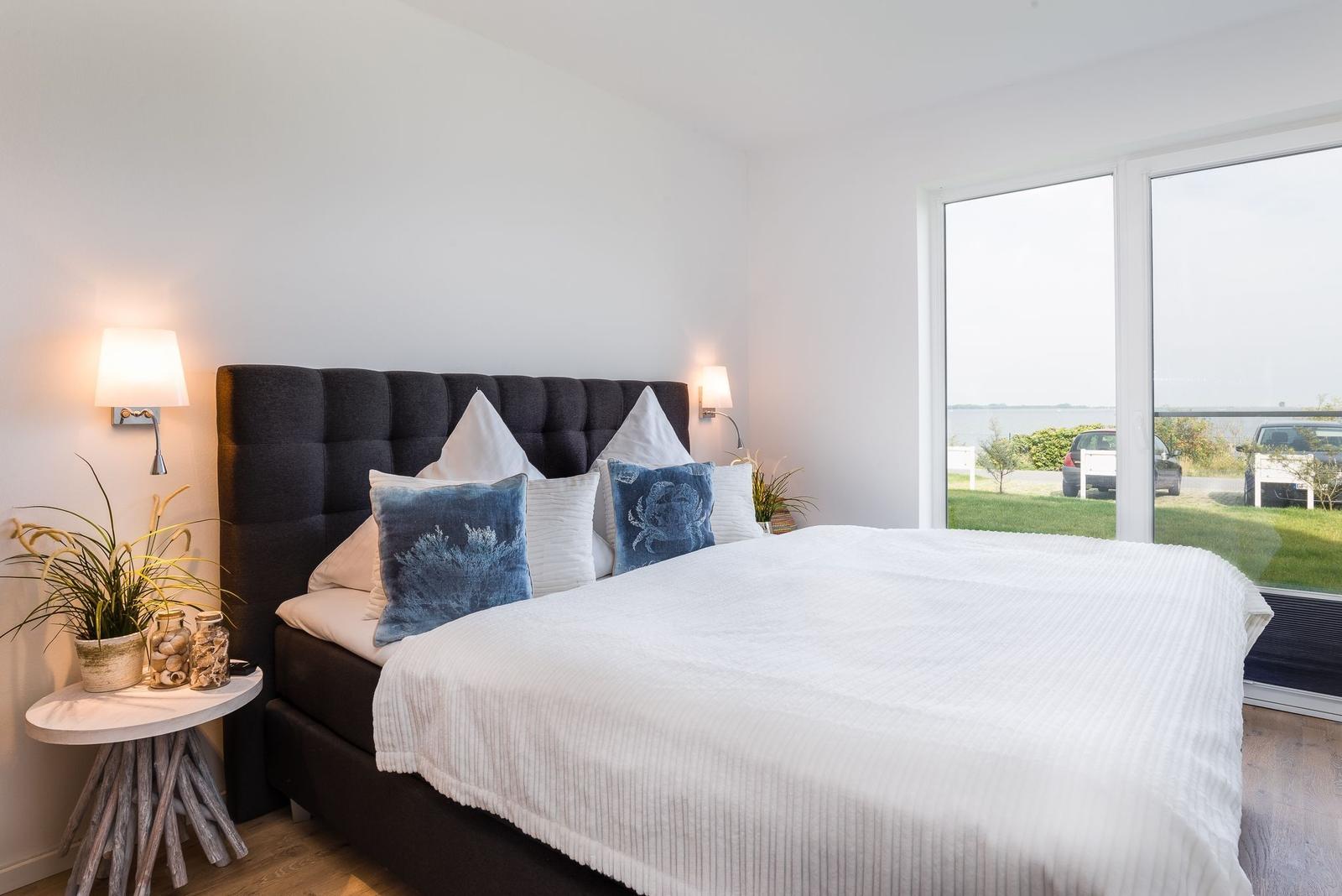 Schlafzimmer mit großem Bett, Fensterblick auf Wasser und Pflanzen auf Nachttisch.