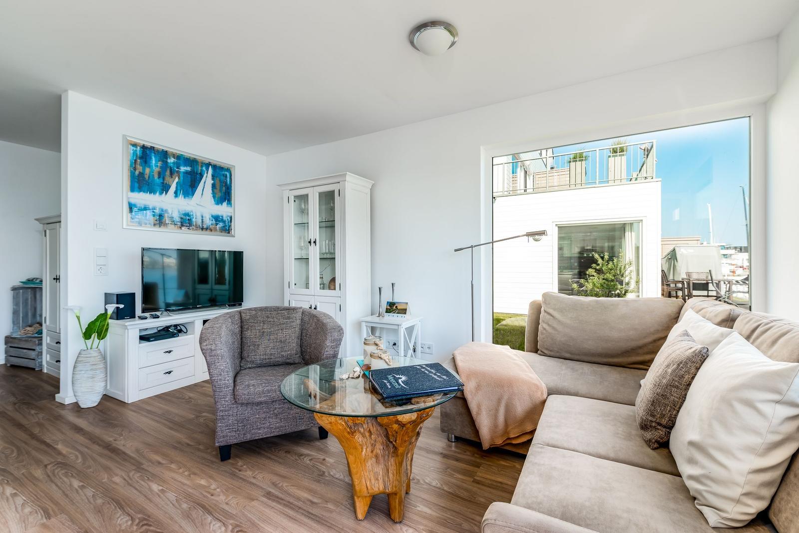 Living room with sofa, armchair, glass table, and TV. Large windows with balcony view.