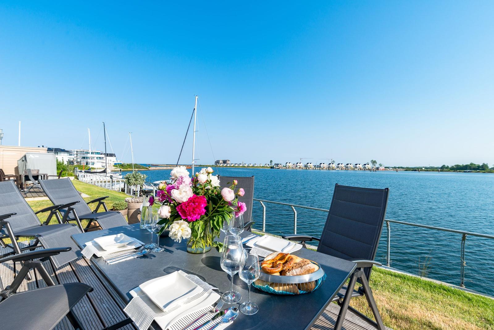 Table with flowers and food on terrace with view of water and sailboats.
