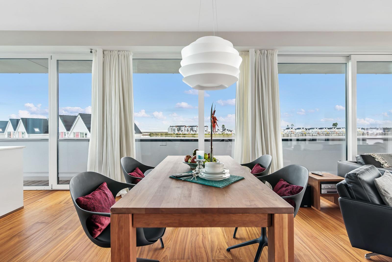 Essbereich mit Holztisch, schwarzen Stühlen und roten Kissen. Große Fenster mit Blick auf Wohngebiet.