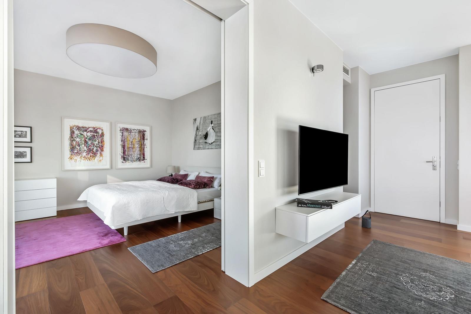 Schlafzimmer mit Bett, Wandbildern und Teppich. Wohnbereich mit Fernseher und Wandregal.