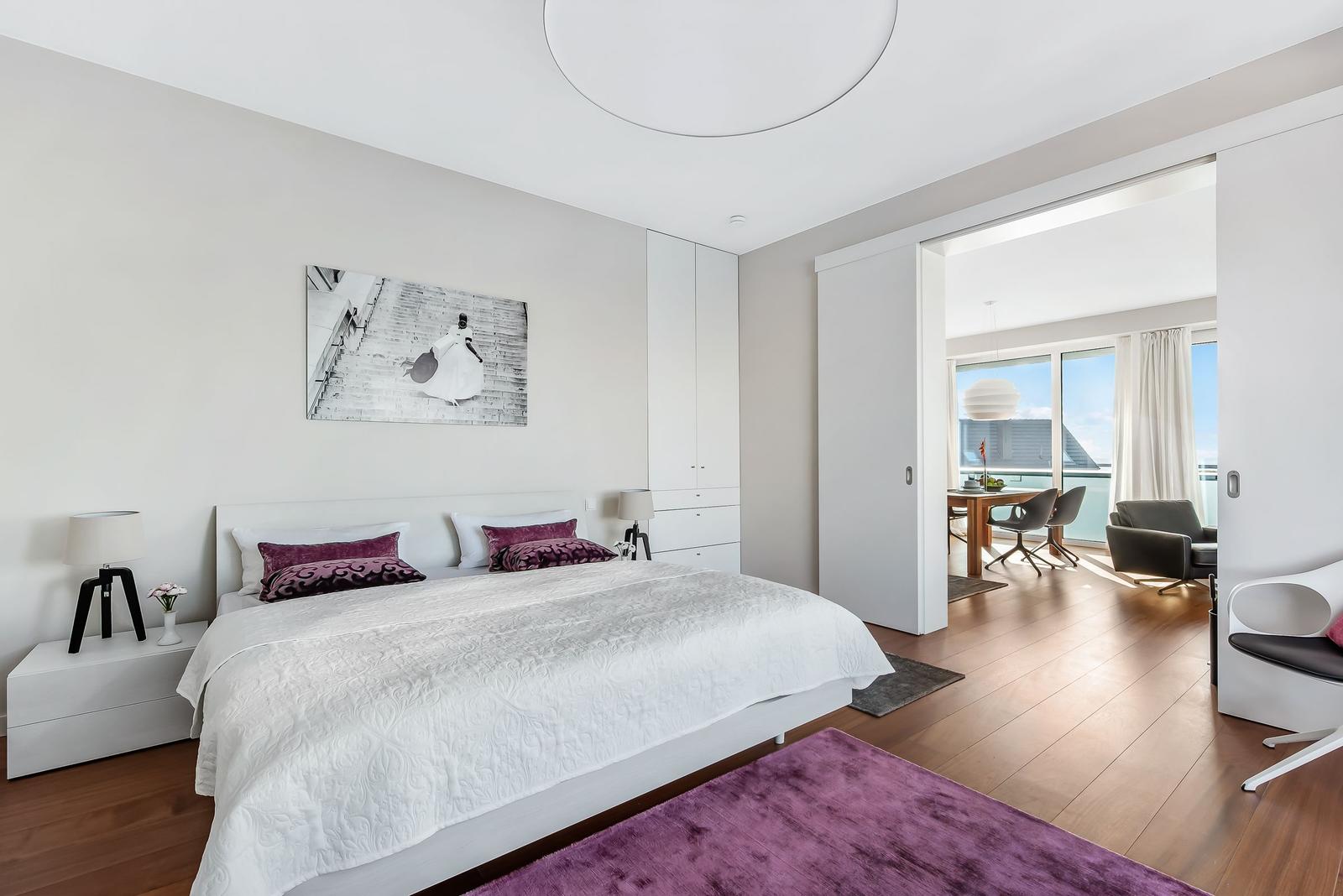 Schlafzimmer mit Bett, Wandbild und Blick in Wohnbereich mit Tisch und Sitzgelegenheiten.