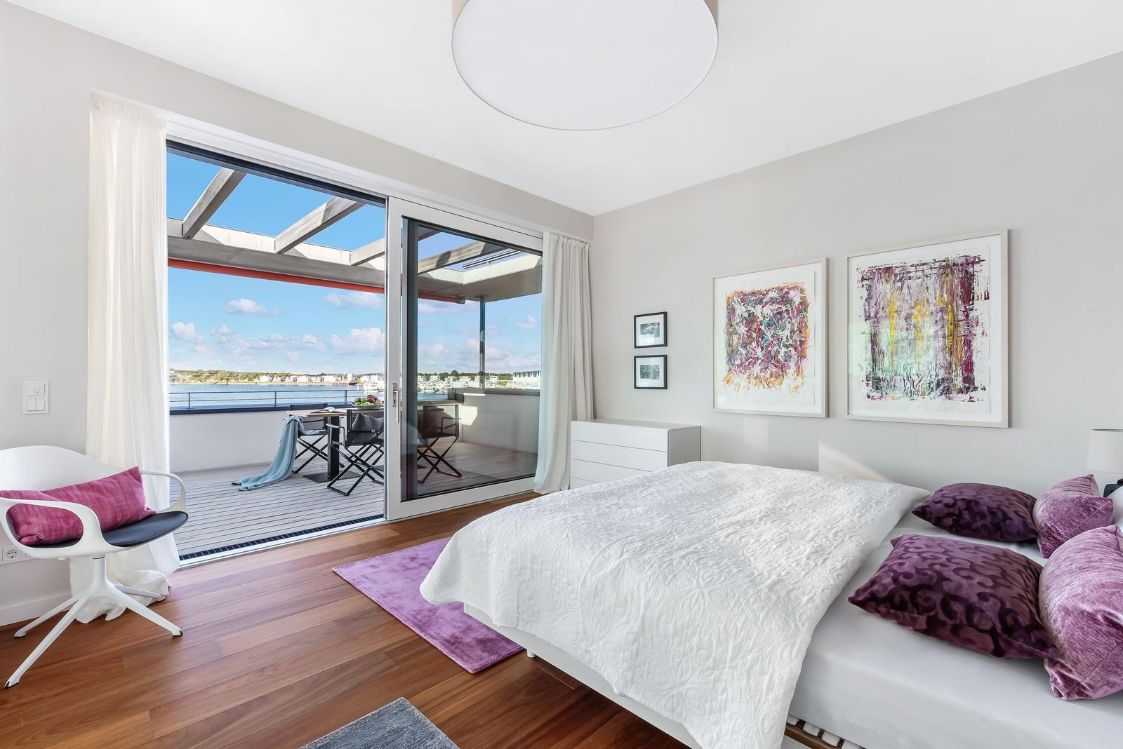 Schlafzimmer mit Balkon und Blick auf Wasser. Bett, Kunstwerke und Holzboden.