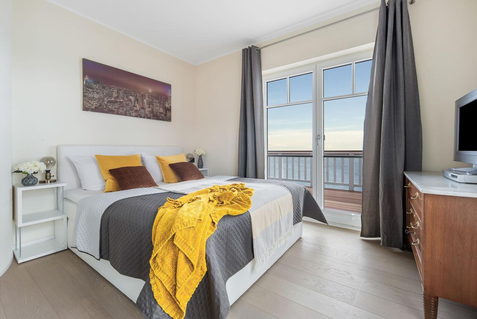 Schlafzimmer mit Blick auf die See, Bett mit Kissen und Decke, Fenster zur Terrasse.