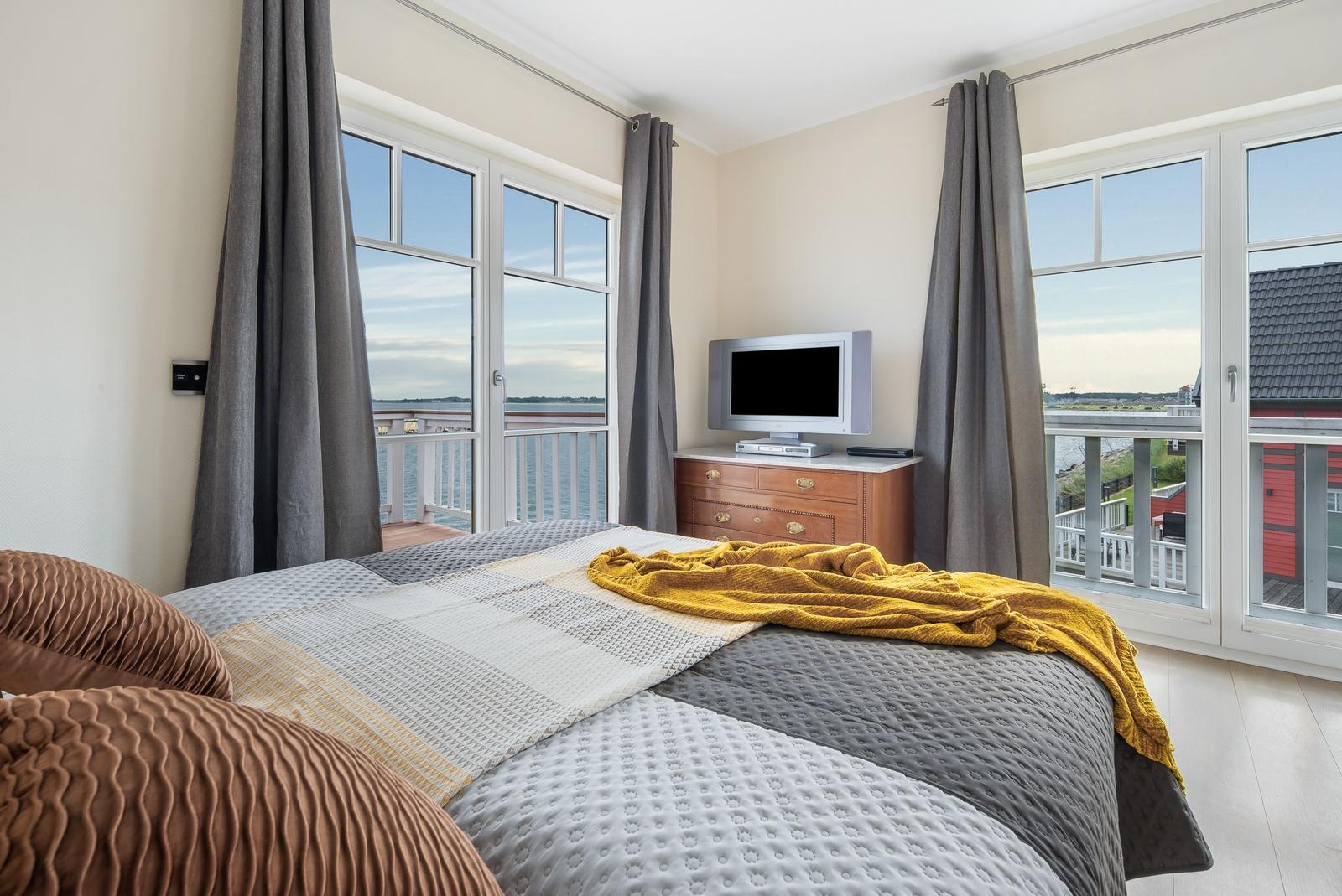 Schlafzimmer mit Blick auf Wasser und Balkon. Bett mit Decke, Fernseher und Schrank.