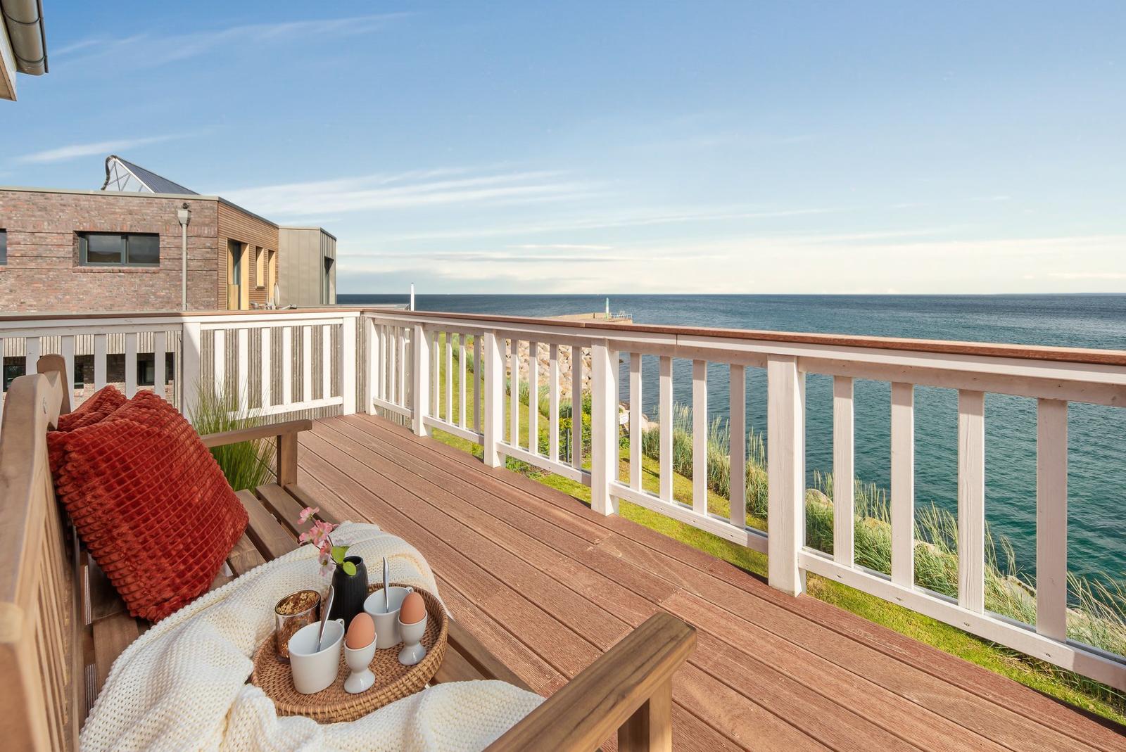Terrasse mit Blick auf das Meer, Holzbank, Kissen, Tisch mit Frühstück.