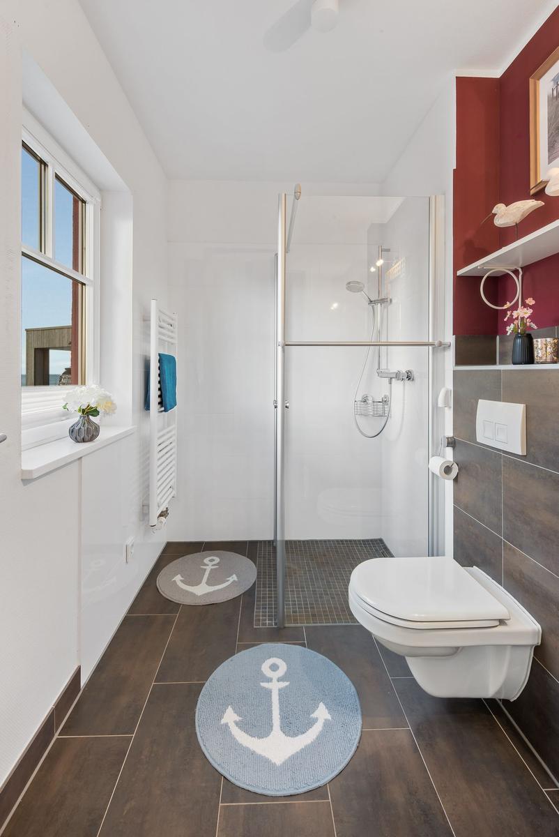 Badezimmer mit Dusche, Toilette und Fenster. Bodenfliesen und zwei runde Teppiche mit Ankern.