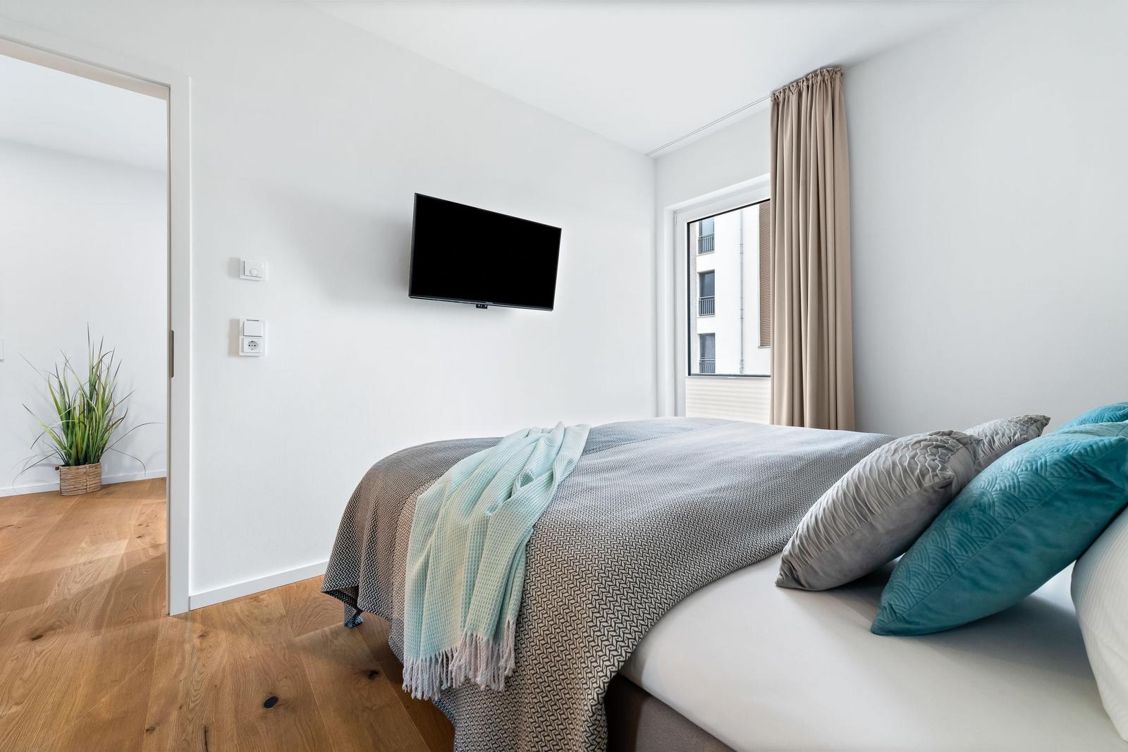 Schlafzimmer mit Bett, Fernseher und Fenster mit Blick auf Gebäude.