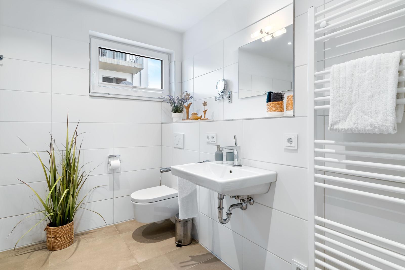 Badezimmer mit Toilette, Waschbecken und Heizkörper. Fenster, Pflanze und Spiegel sichtbar.