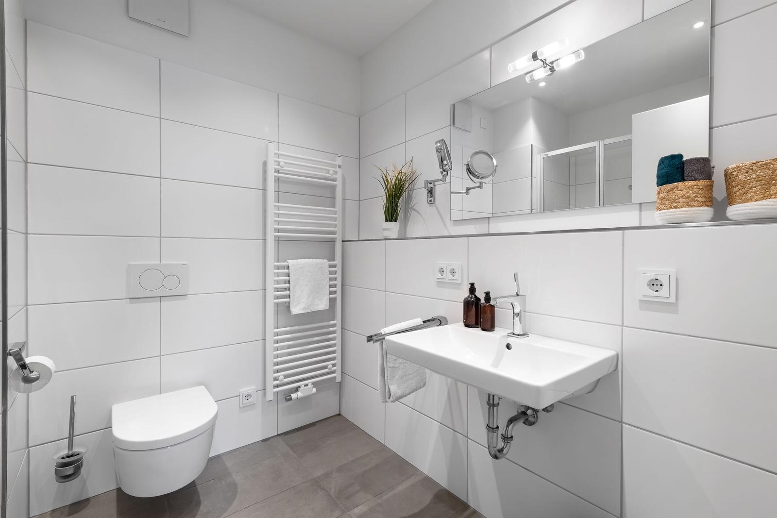 Badezimmer mit Toilette, Waschbecken und Heizkörper. Weiße Fliesen und Spiegel.