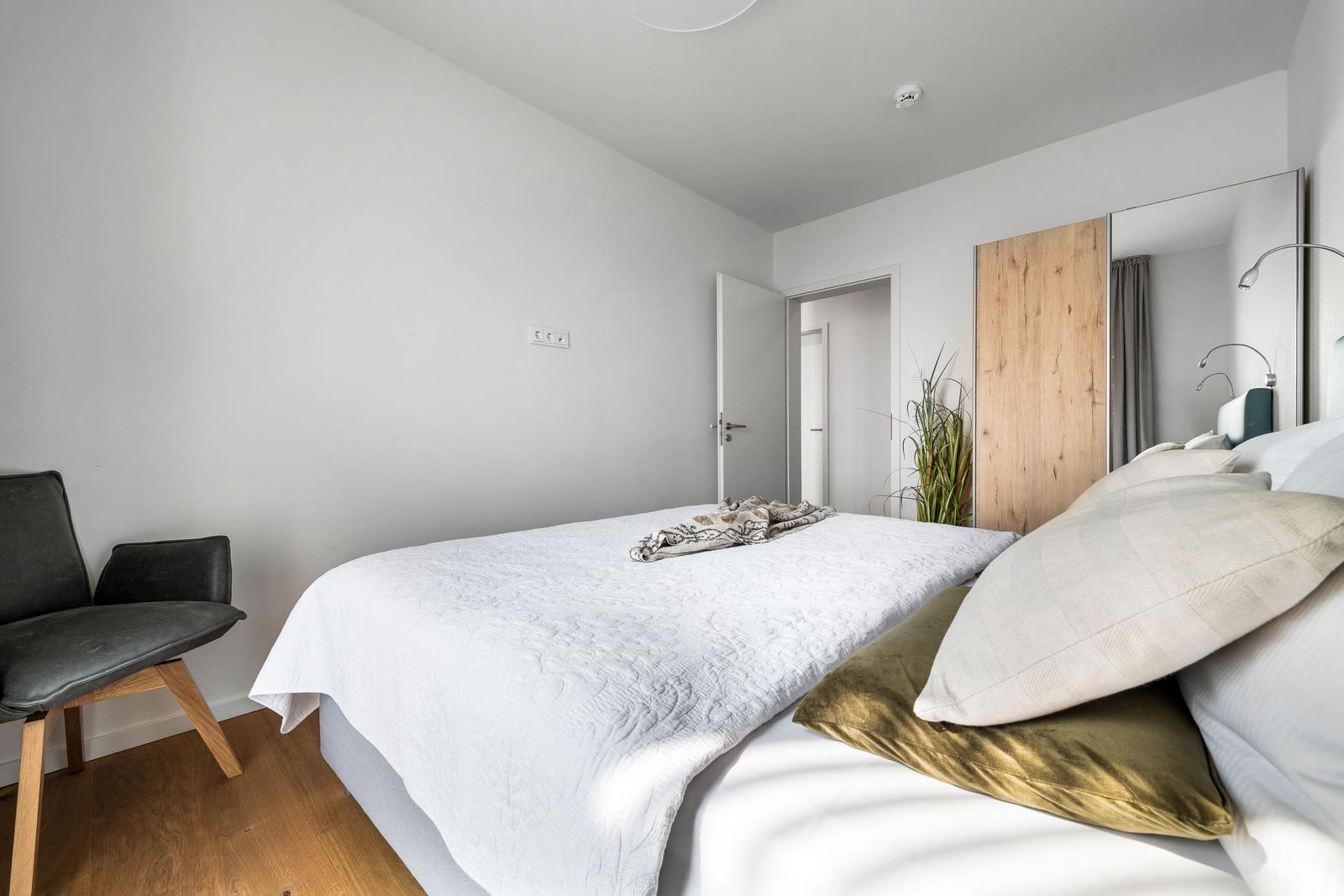 Schlafzimmer mit Bett, Stuhl und Spiegel. Holzboden und weiße Wände.