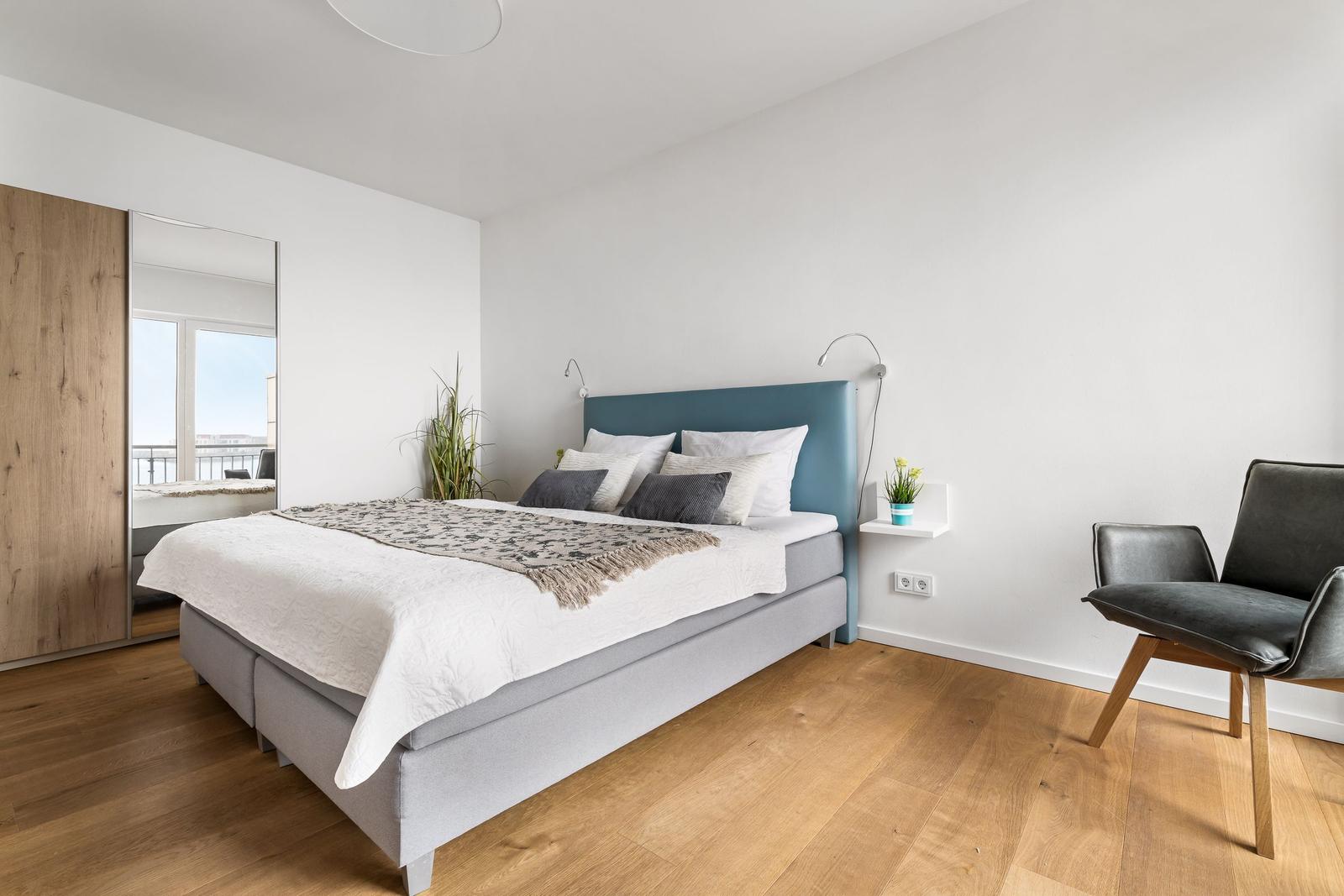 Schlafzimmer mit Bett, Stuhl und Spiegelschrank. Holzboden und blaues Kopfteil.