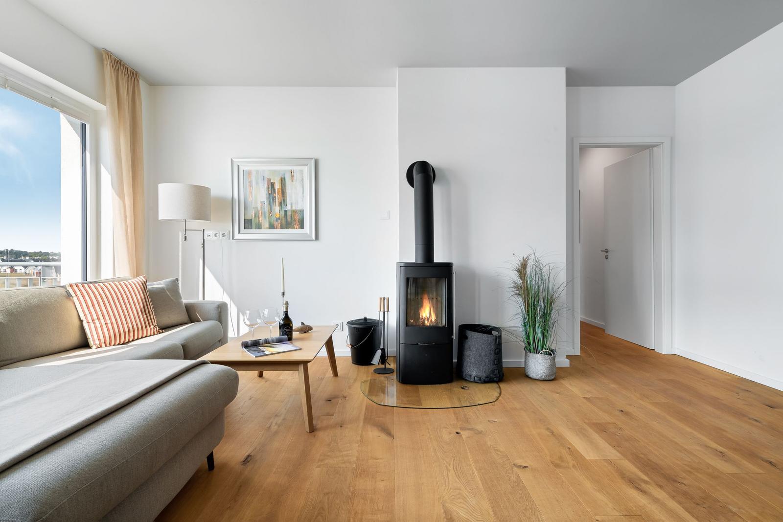 Wohnzimmer mit Holzofen, Sofa und großem Fenster. Holzboden und weiße Wände.