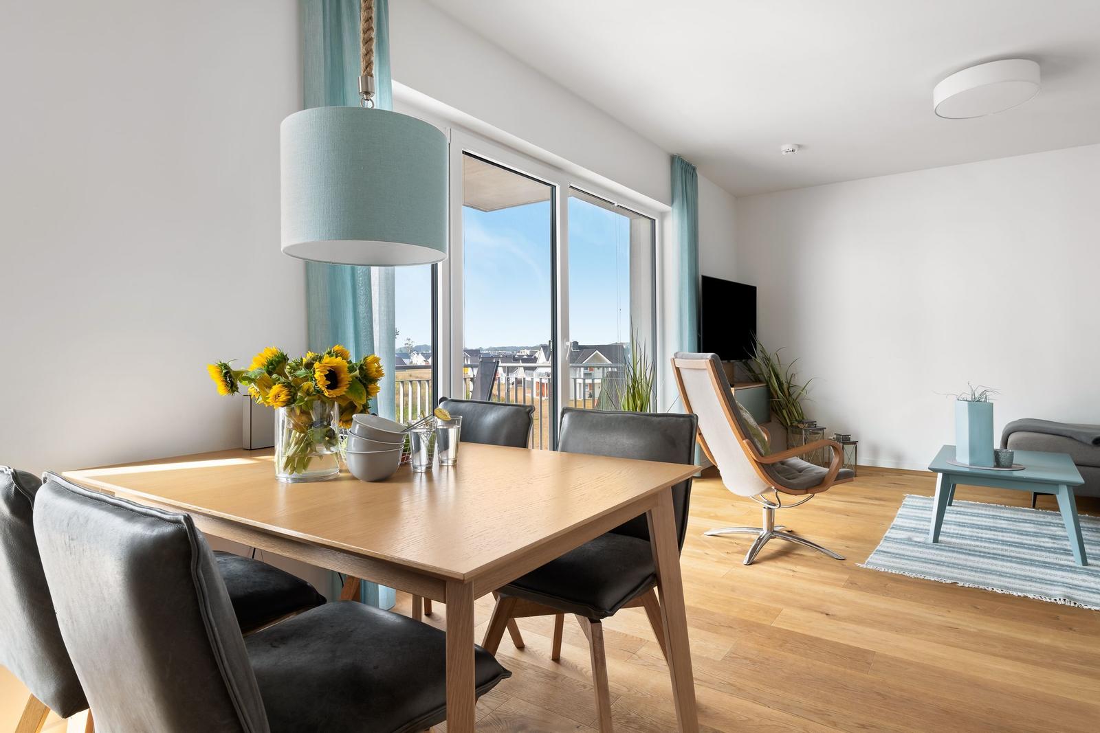 Essbereich mit Tisch, Stühlen und Sonnenblumen. Wohnbereich mit Sessel und TV. Holzboden und große Fenster.
