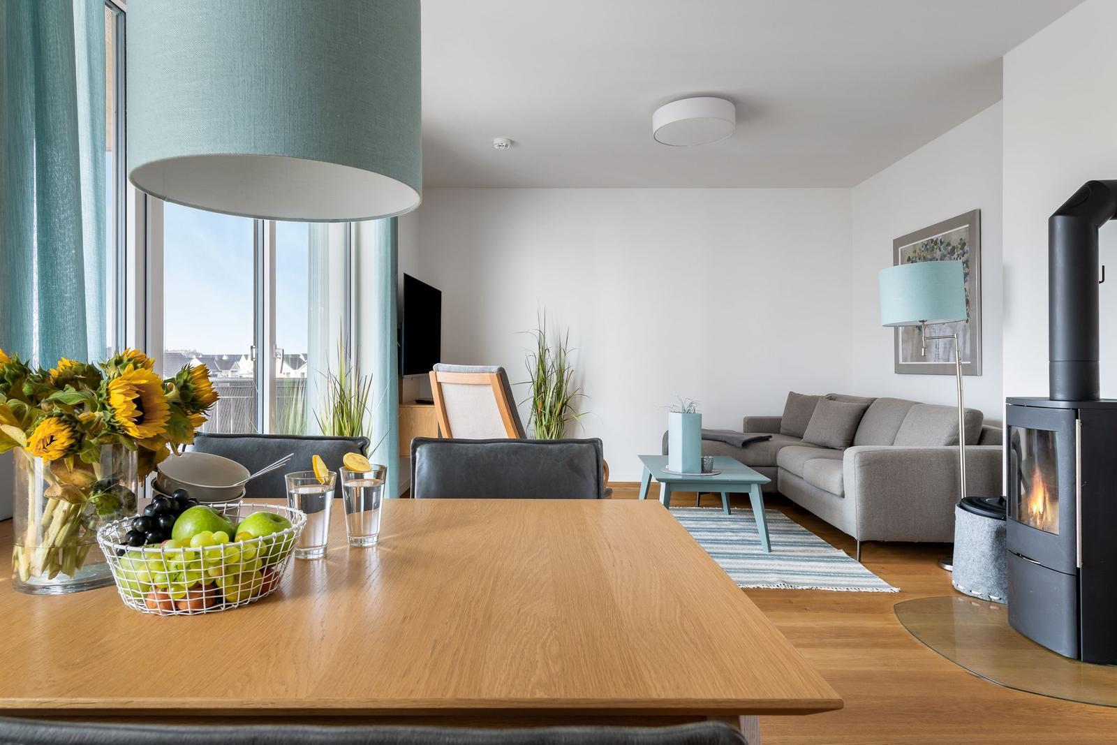 Ein Essbereich mit Holztisch und Obst. Im Hintergrund ein Wohnzimmer mit Sofa und Kamin.