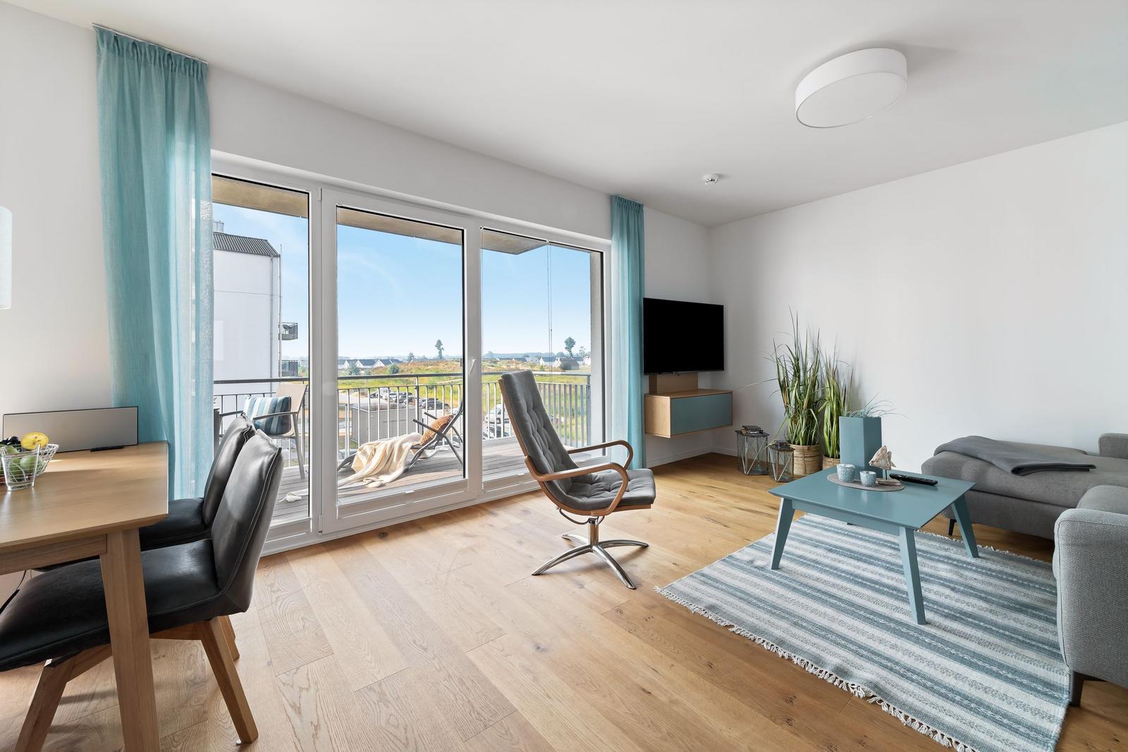 Wohnbereich mit Balkon, Sofa, Sessel und TV. Holzboden und blaue Akzente.