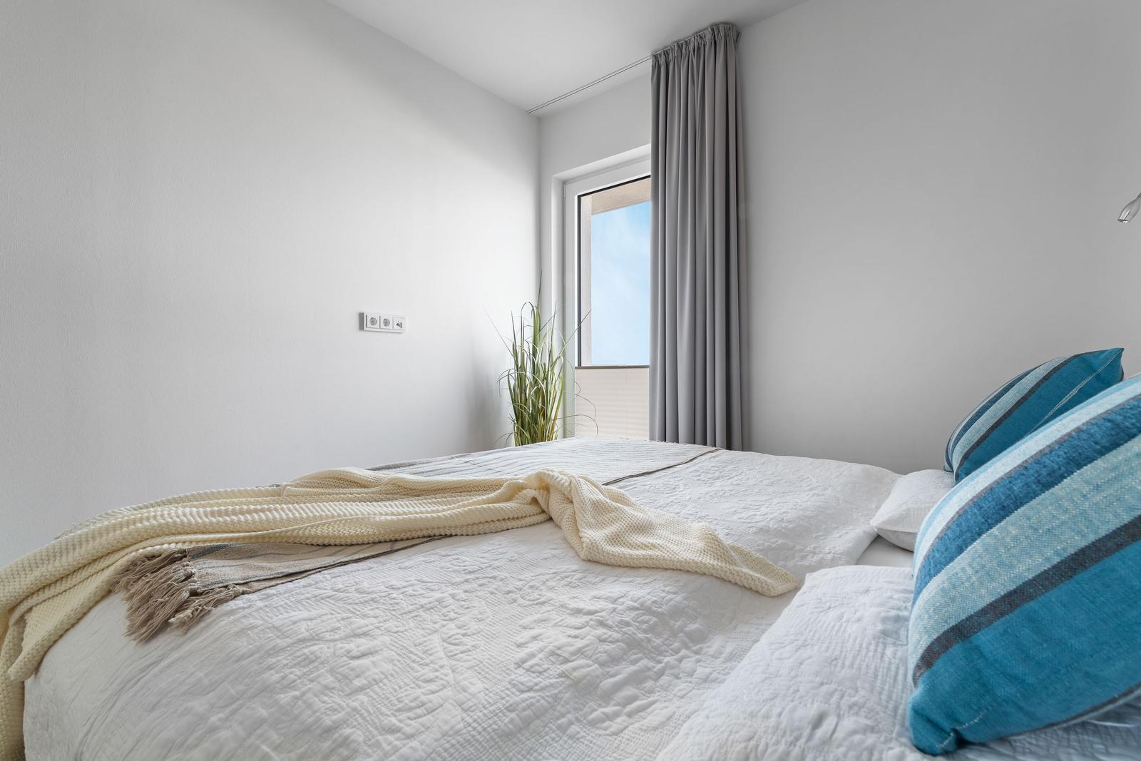 Schlafzimmer mit Bett, blauen Kissen und Fenster mit grauen Vorhängen.