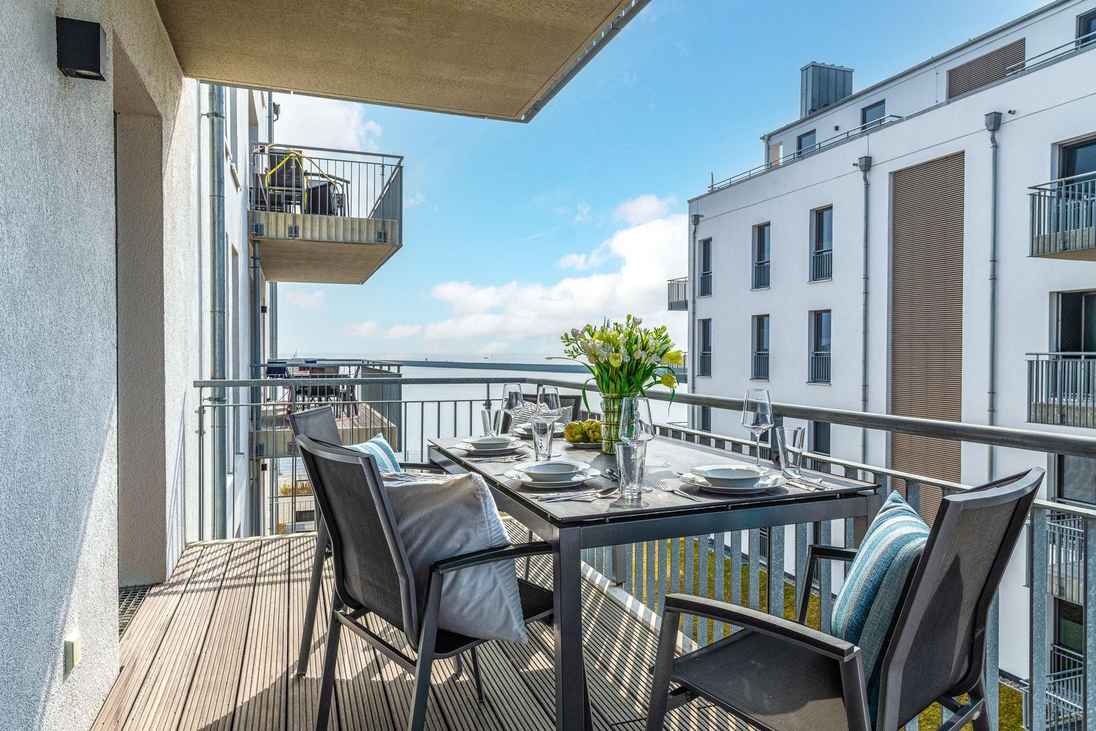 Terrasse mit Tisch, Stühlen und Blick auf Wasser und Gebäude.
