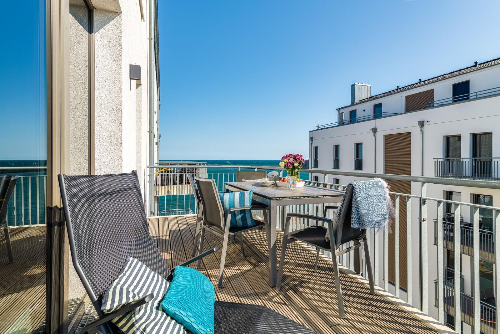 Terrace with table, chairs, and sea view. Blue cushions and flowers on the table.