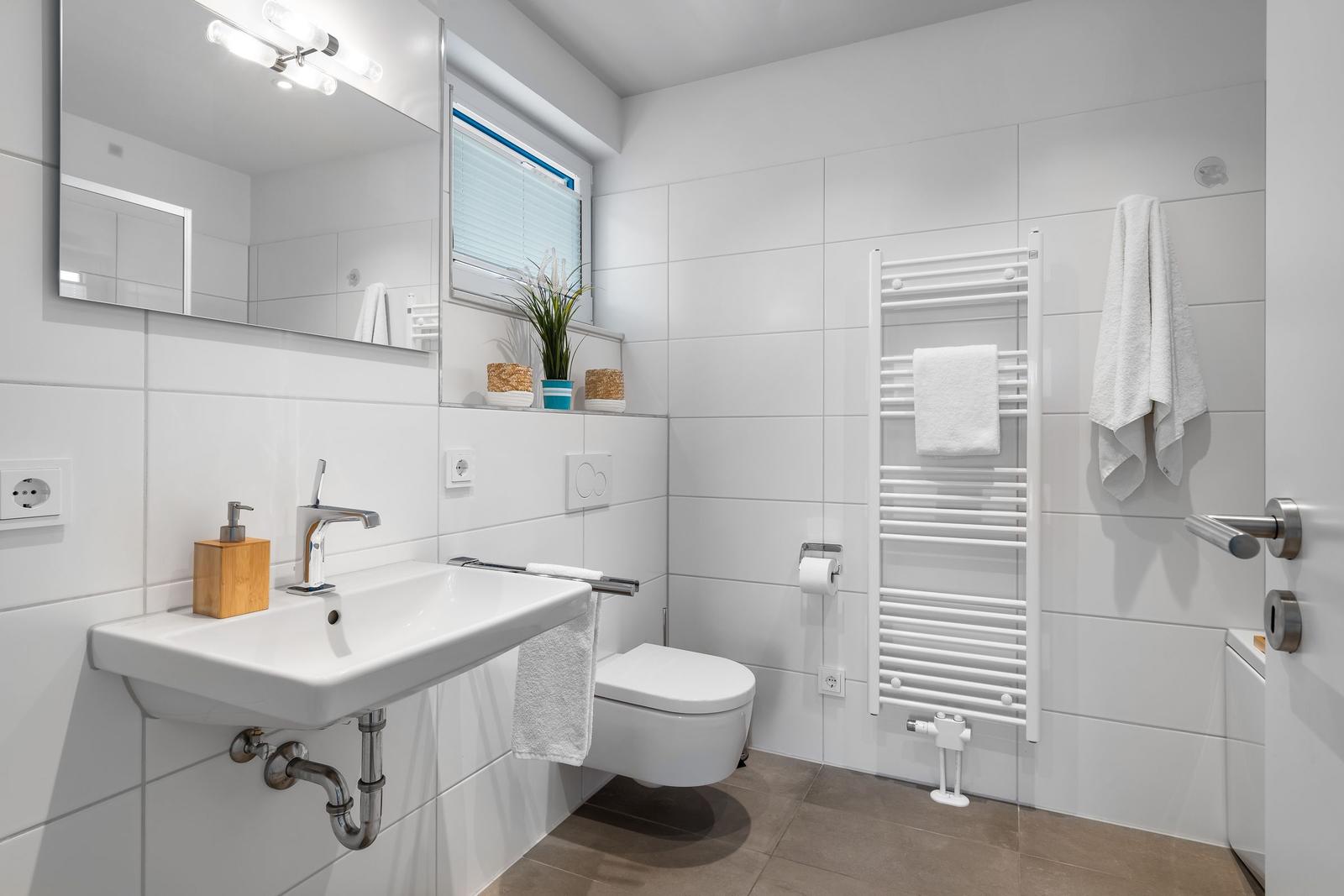 Bathroom with sink, toilet, and heater. White tiles and window with blinds.