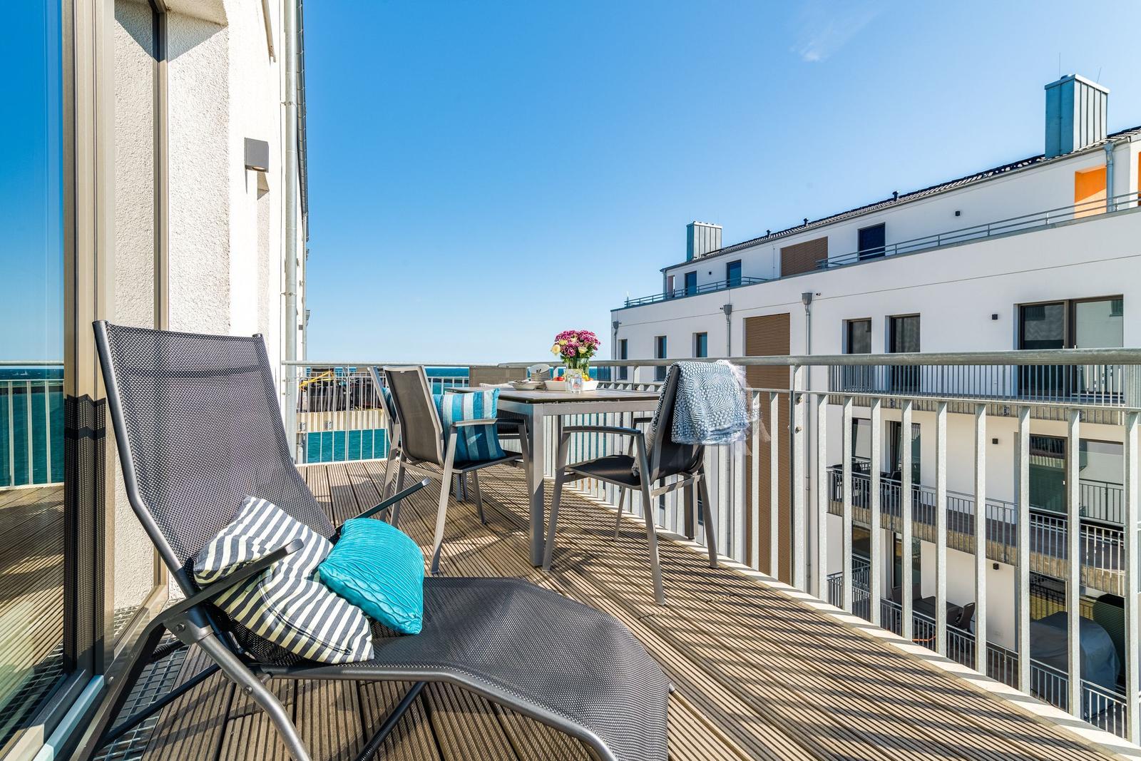 Balcony with lounge chair, table, and chairs. View of buildings and sea.