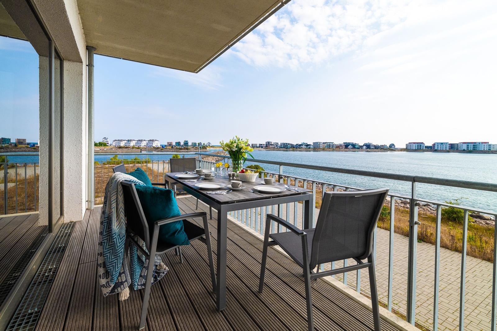 Terrasse mit Tisch und Stühlen, Blick auf Wasser und gegenüberliegende Gebäude.