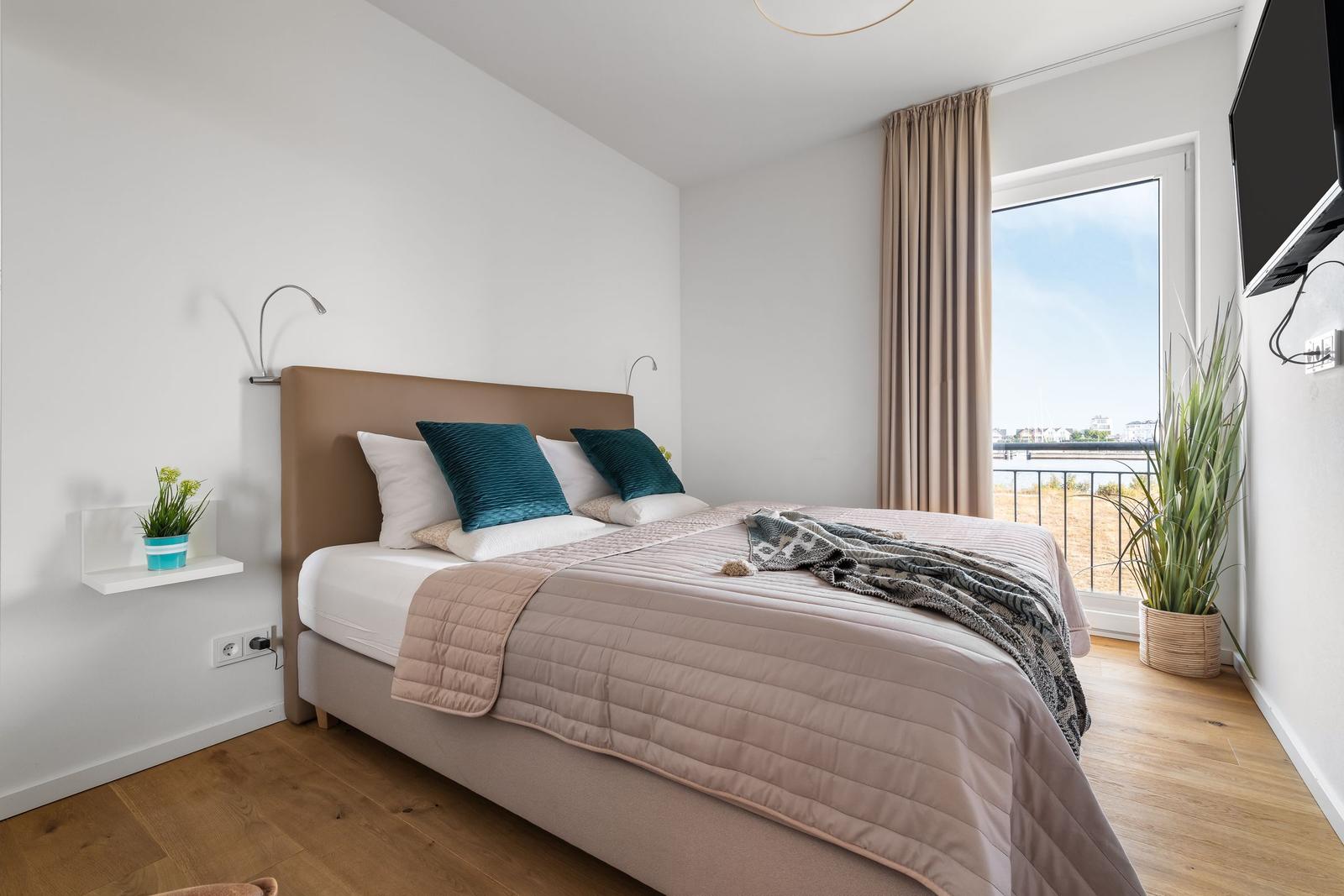 Schlafzimmer mit Bett, Fenster und Pflanzen. Holzboden und Wandhalterungen.