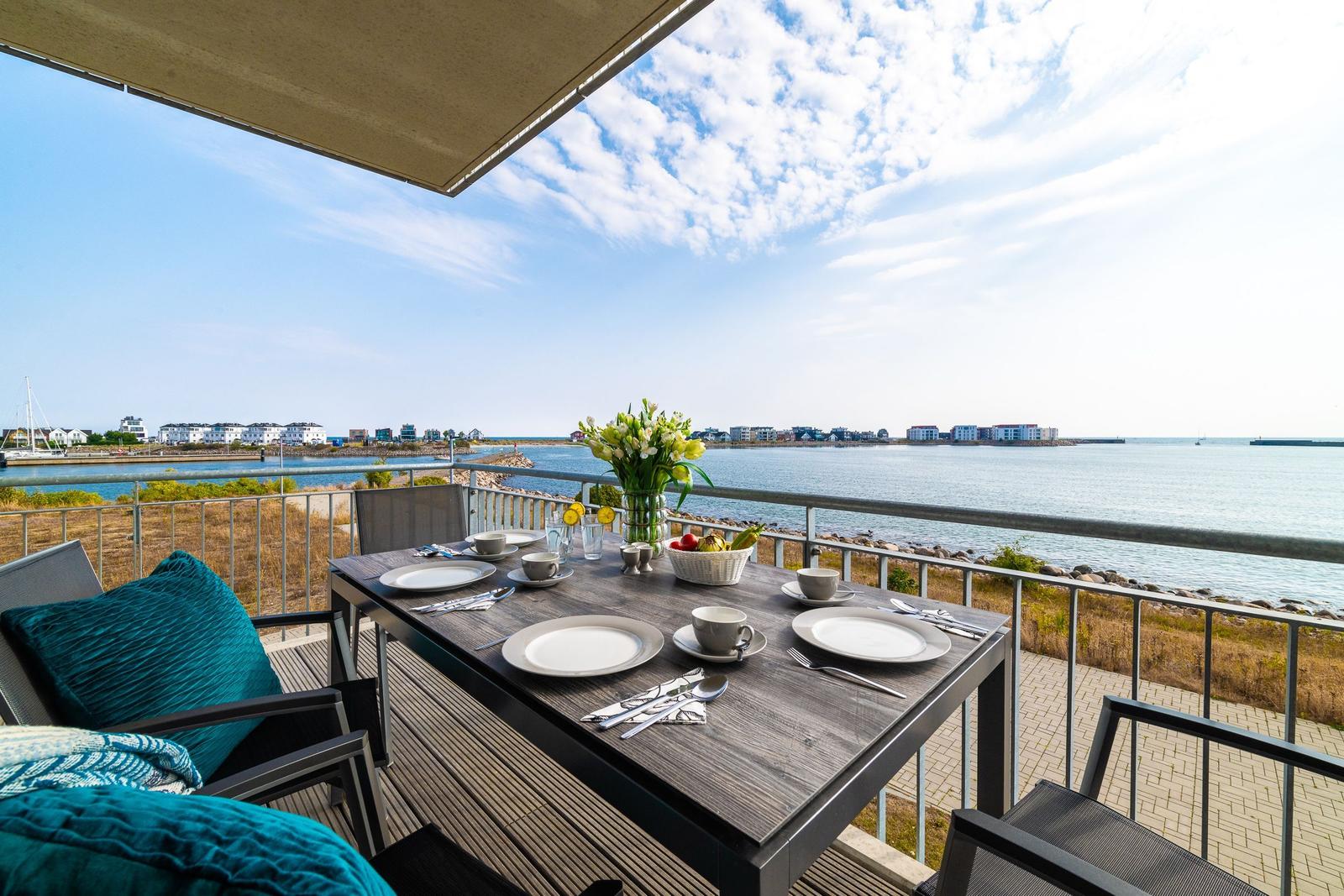 Terrasse mit Tisch, Stühlen und Meerblick. Blumen, Obst und Geschirr sind darauf platziert.