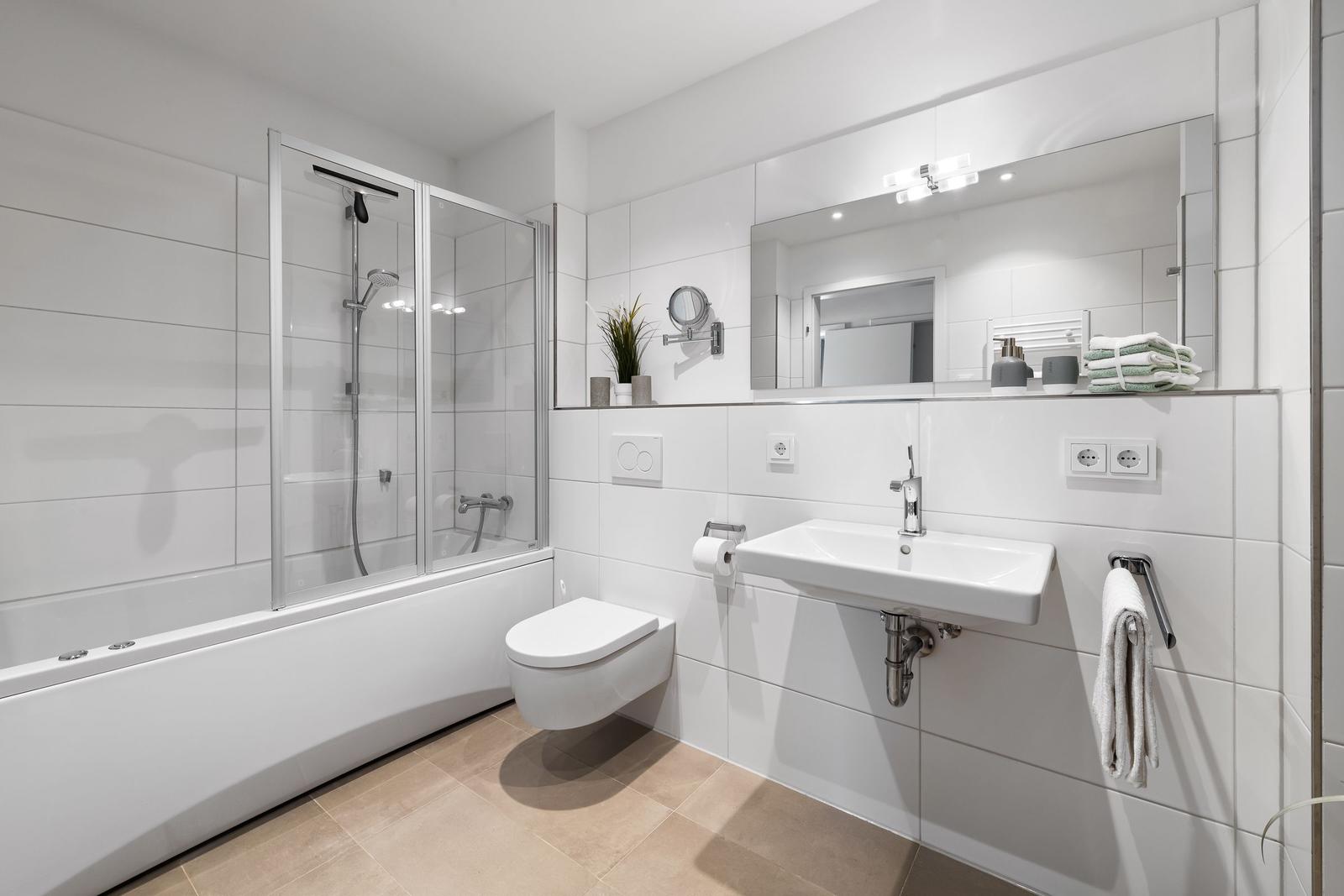 Badezimmer mit Dusche, Waschbecken und Toilette. Weiße Fliesen und Handtücher.